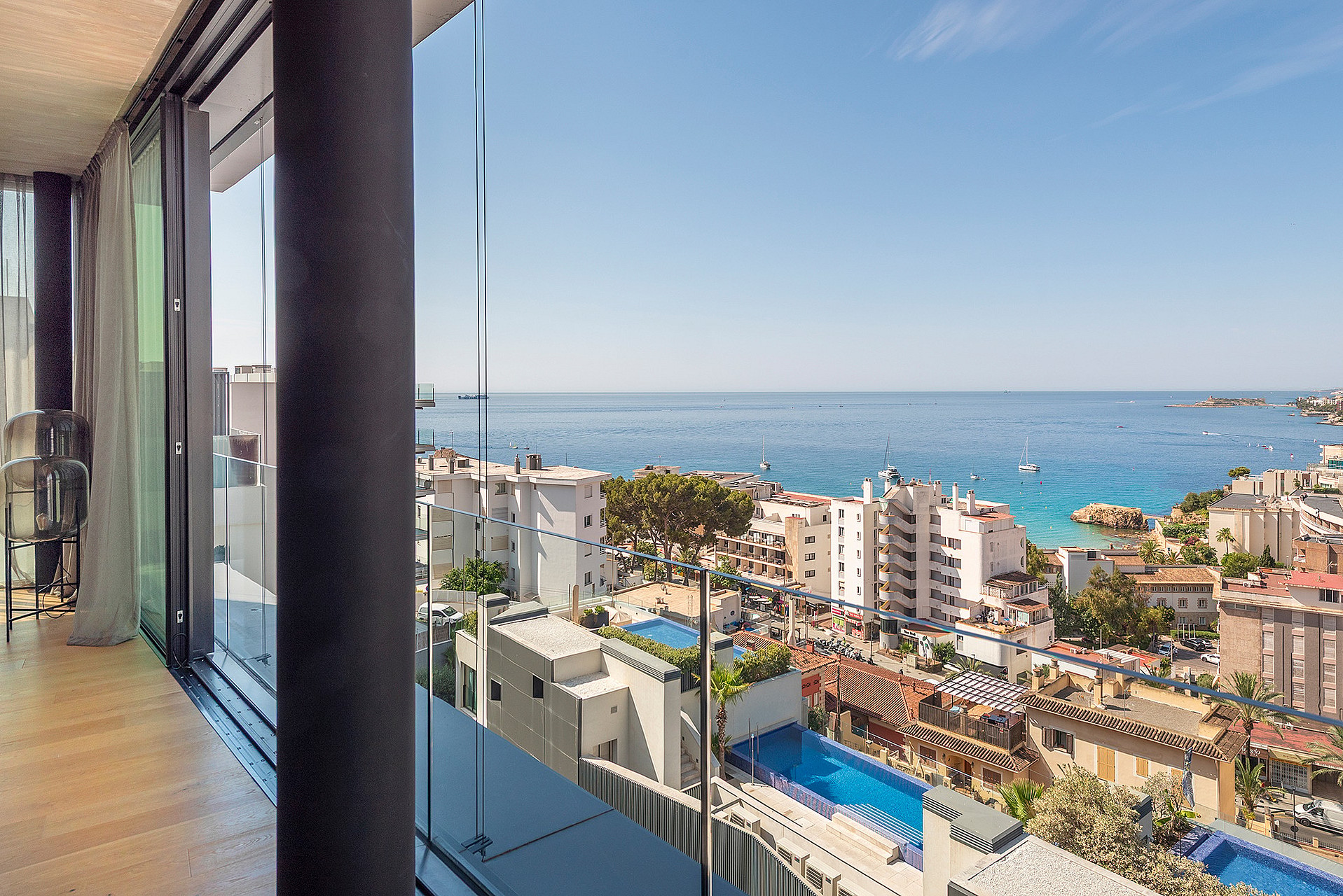 A glass-railed balcony looking out over the urban coastline of Cala Mayor with a swimming pool visible below.