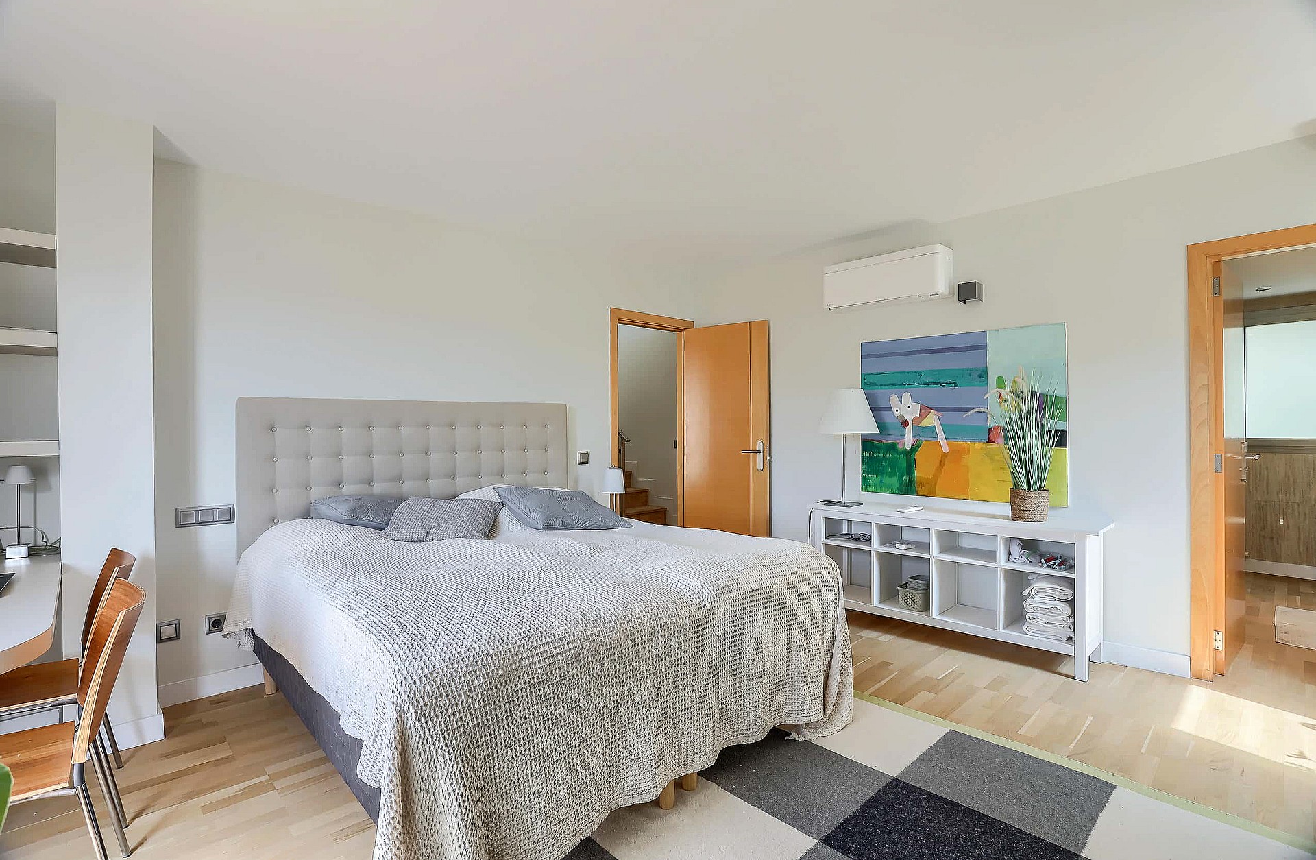 A bright master bedroom with a tufted headboard, light oak flooring, and a modern painting above a white shelving unit.