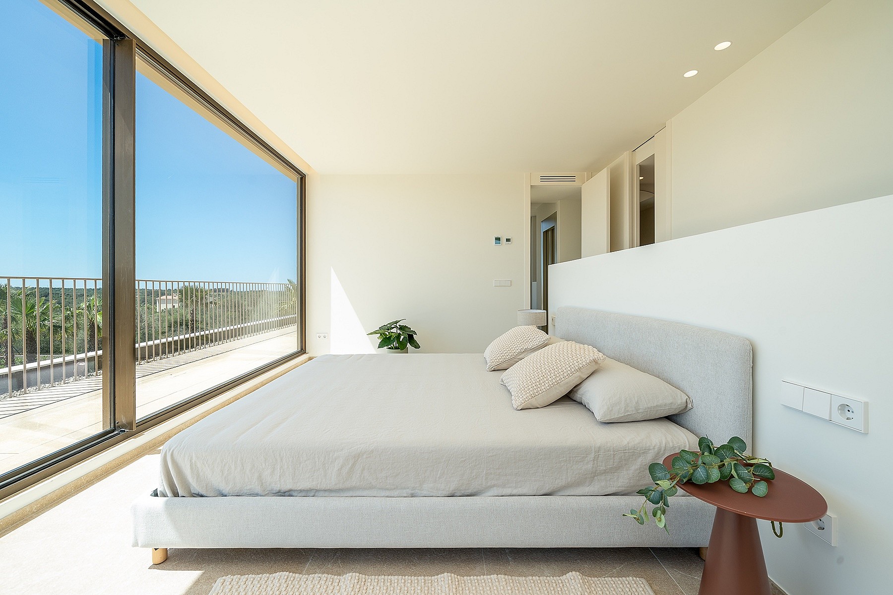 Modern bedroom with a double bed, floor-to-ceiling glass wall, white walls, and view of the exterior landscape.