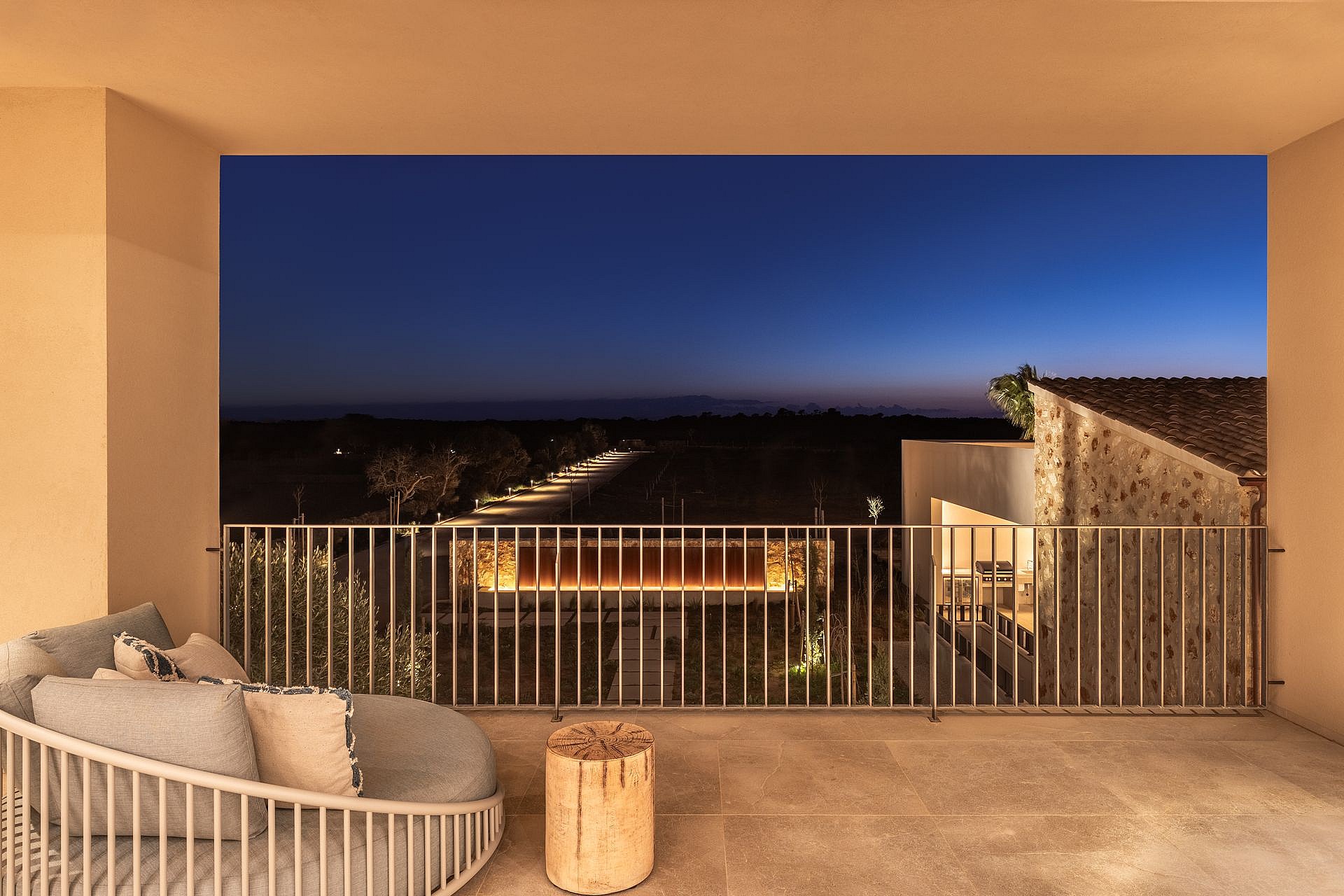A covered terrace with a lounge chair and wooden side table overlooking a landscape at dusk, featuring a metal railing.