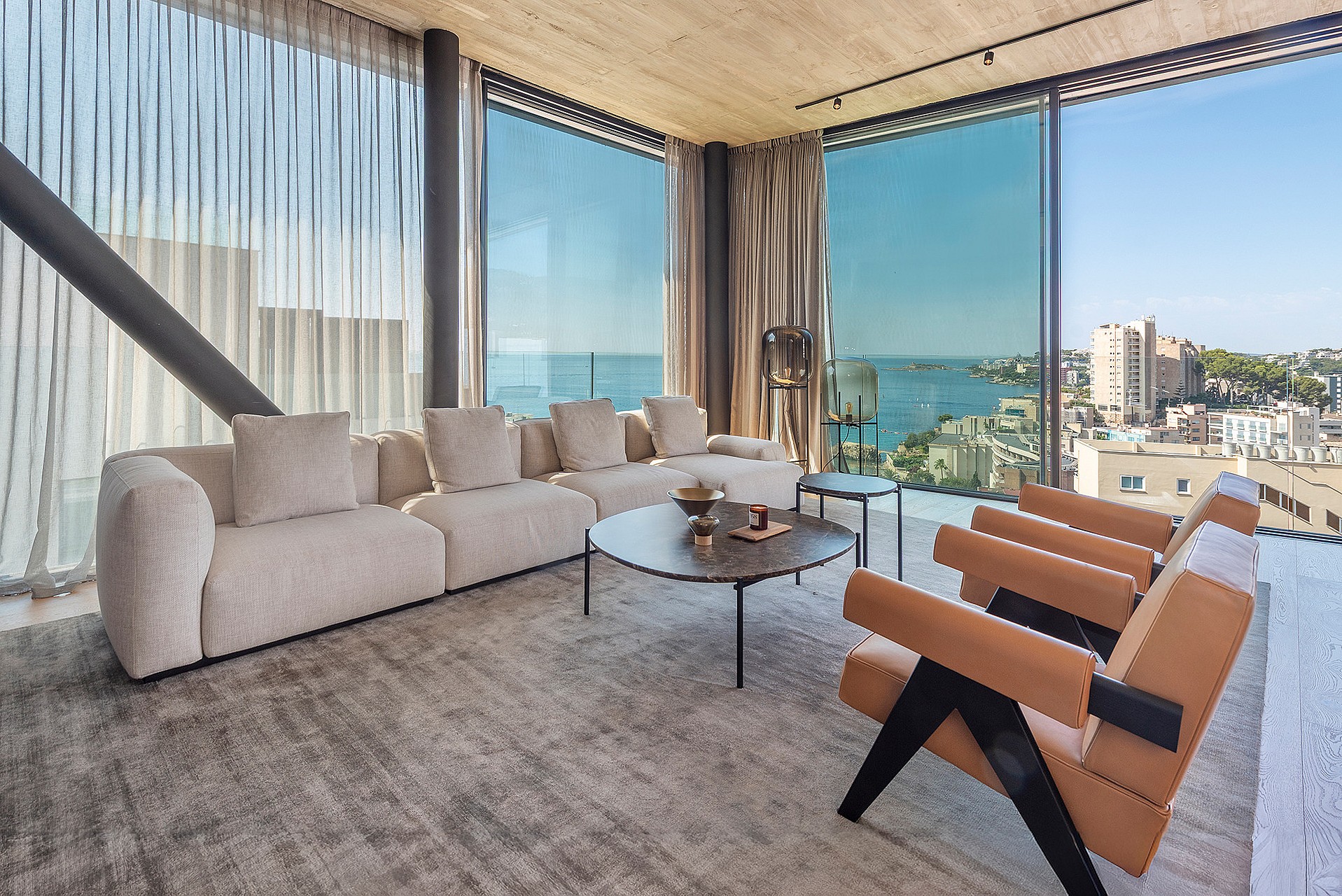 Interior view of a living room featuring modular cream sofa, leather armchairs, and floor-to-ceiling glass walls with sea views.