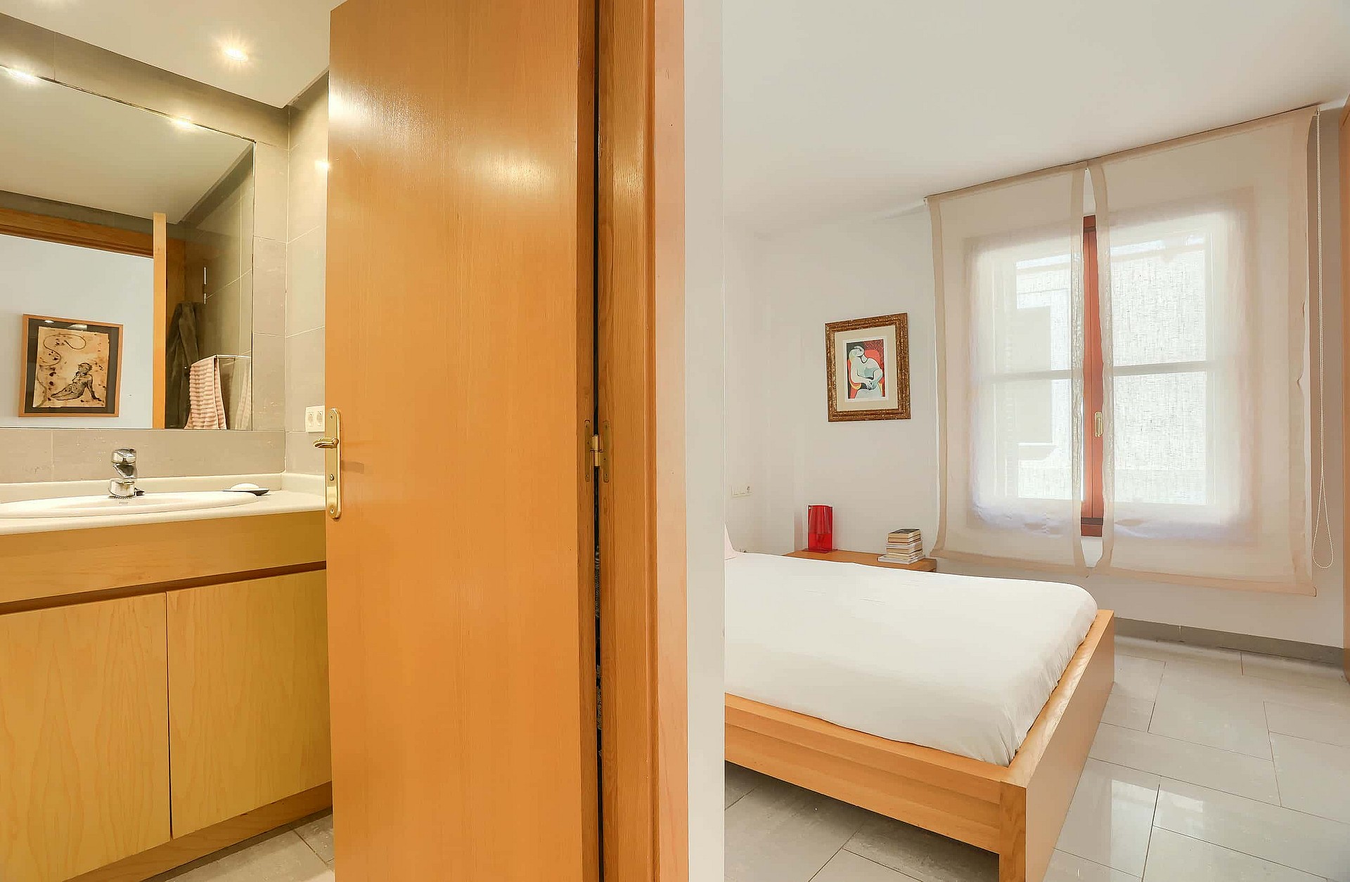 A view through a wooden doorframe showing a bedroom with a bed to the right and a bathroom vanity unit to the left.