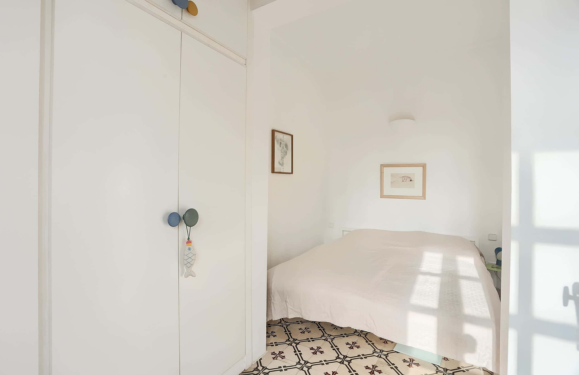 Compact bedroom featuring white walls, a built-in white wardrobe, and decorative patterned floor tiles under a double bed.