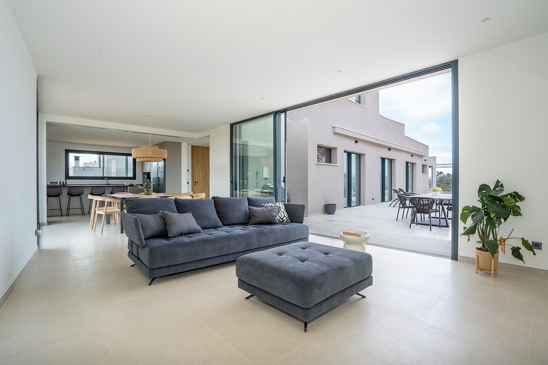A modern living room with a dark grey sofa, light stone flooring, and sliding glass walls opening to an outdoor terrace.
