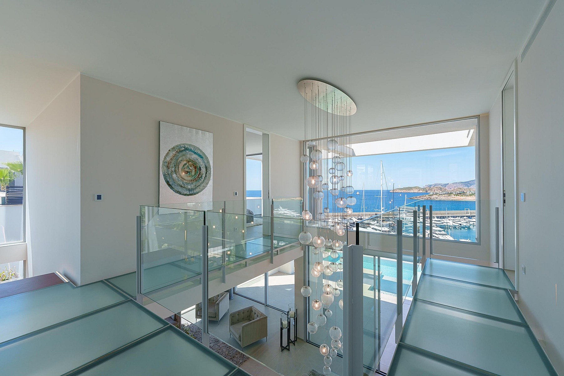 A glass-floored landing overlooks a double-height living space with a large feature chandelier and views of a marina.