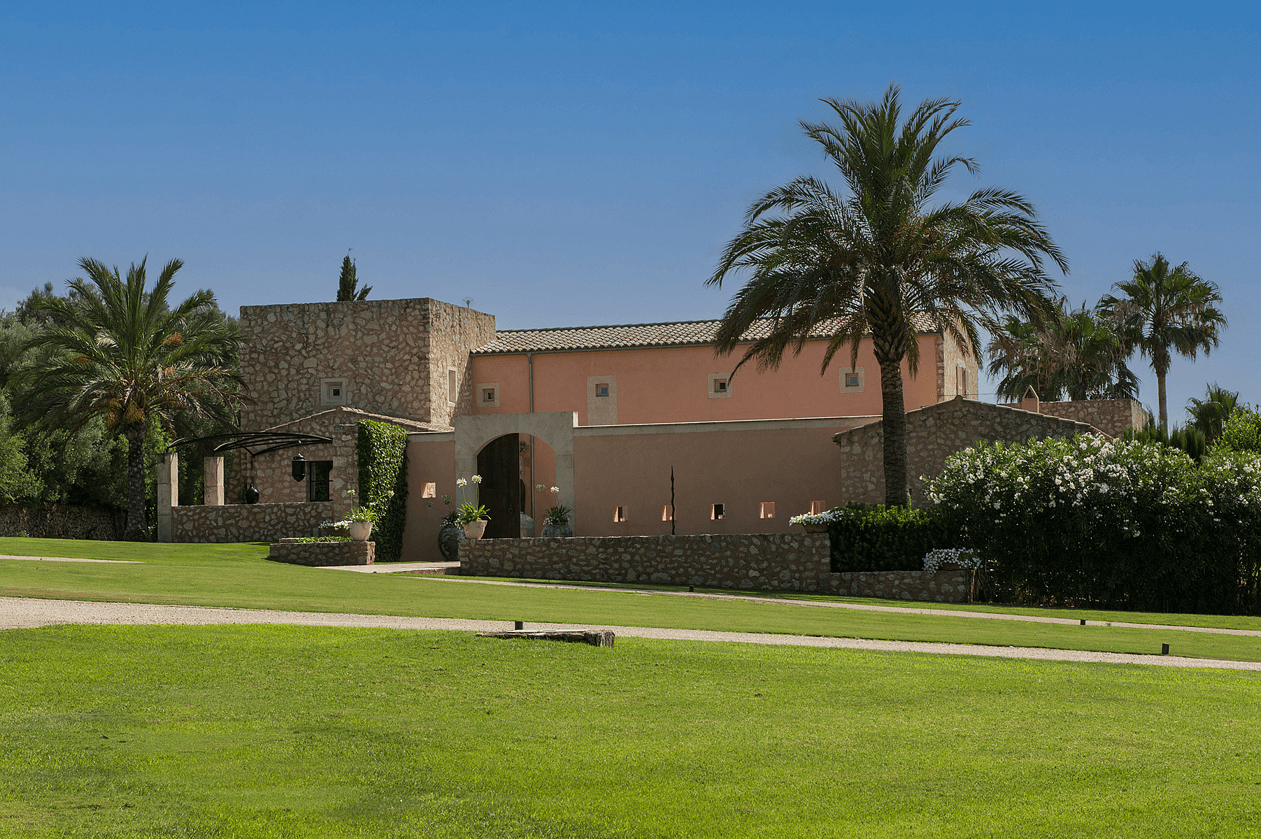 Exterior view of a traditional stone and peach-colored finca with arched entrance, large palm tree, and manicured green lawn.