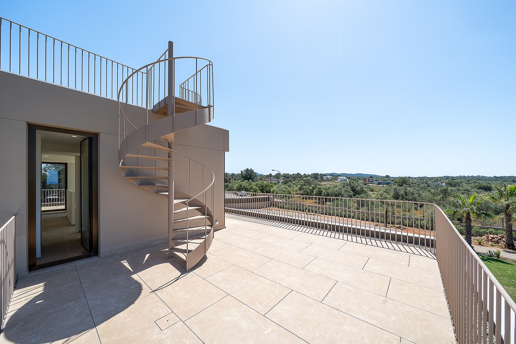 A beige rooftop terrace featuring a metal spiral staircase and minimalist metal railings, overlooking a rural landscape.