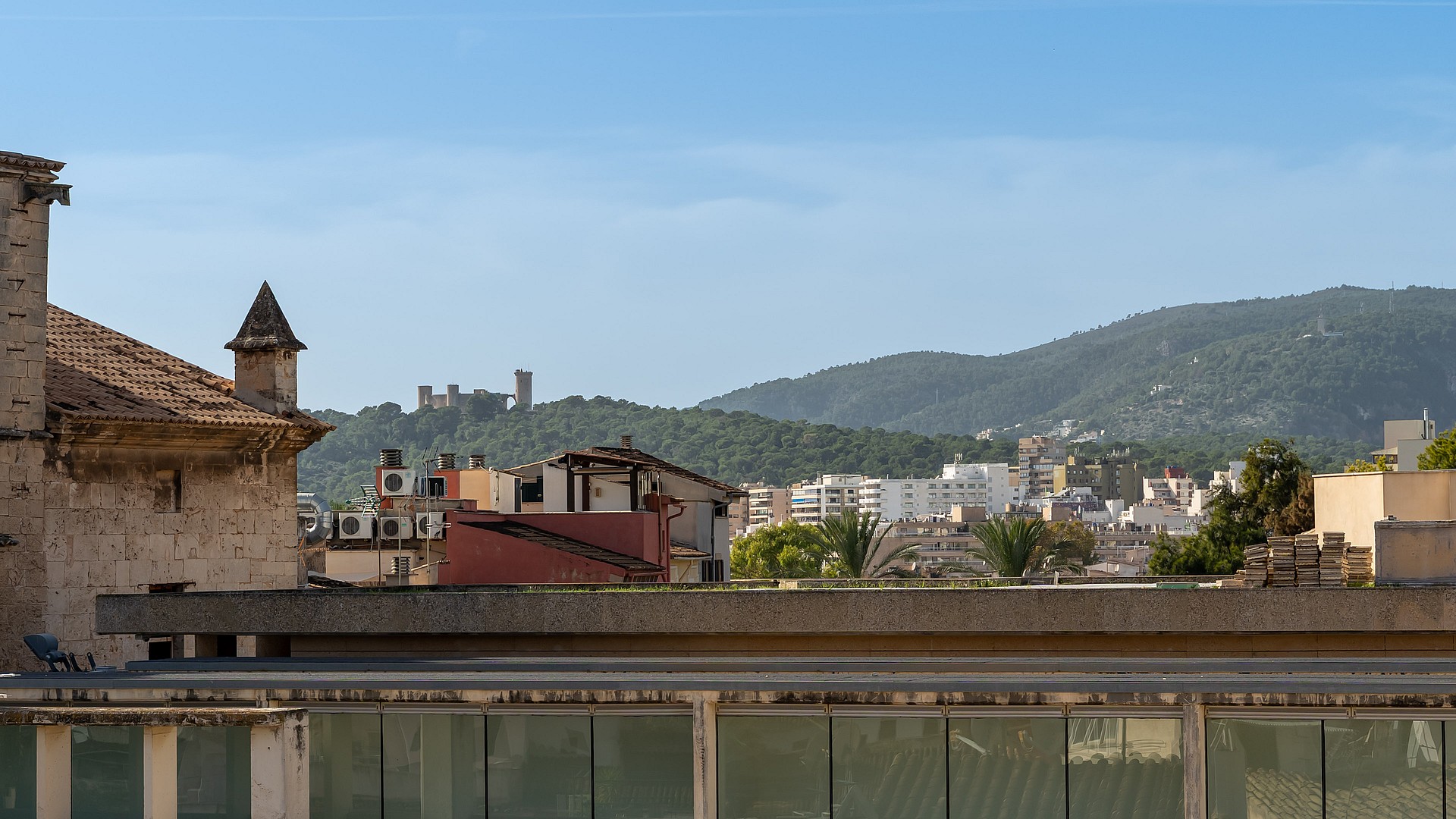 View from a rooftop terrace overlooking historic Palma rooftops and Bellver Castle towards the Tramuntana mountains.
