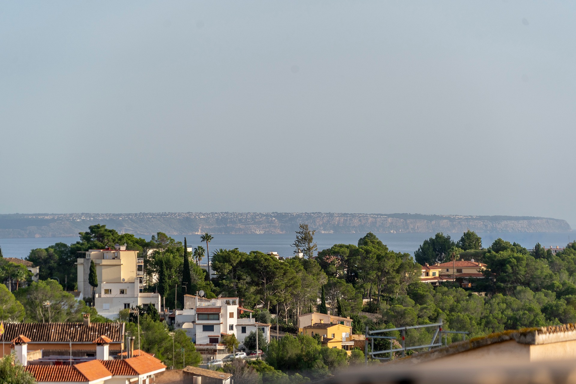 A wide landscape view looking out over trees and Mediterranean style houses towards a distant coastline and sea.