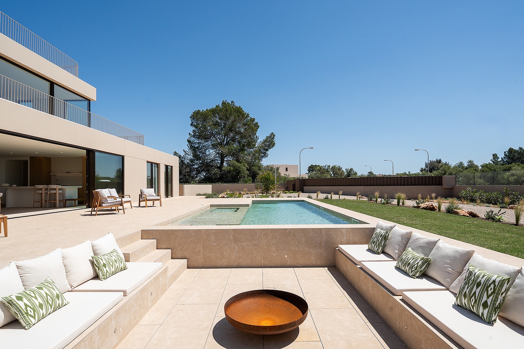 A sunken outdoor seating area with stone benches and white cushions faces a rectangular swimming pool under a blue sky.