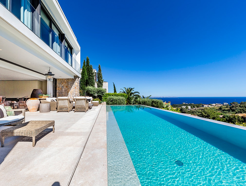 En infinity-pool med turkost vatten mot horisonten, intill en bred stenterrass med lounge-möbler.