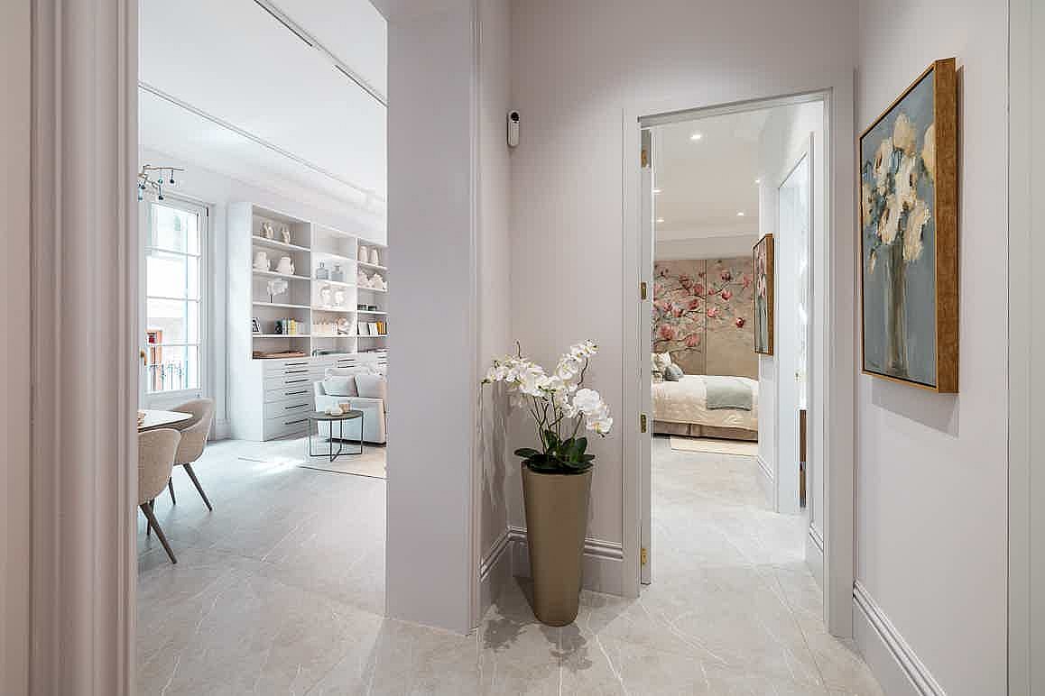 A bright hallway with white walls and stone flooring connecting a bedroom to a living area, featuring a tall plant pot.