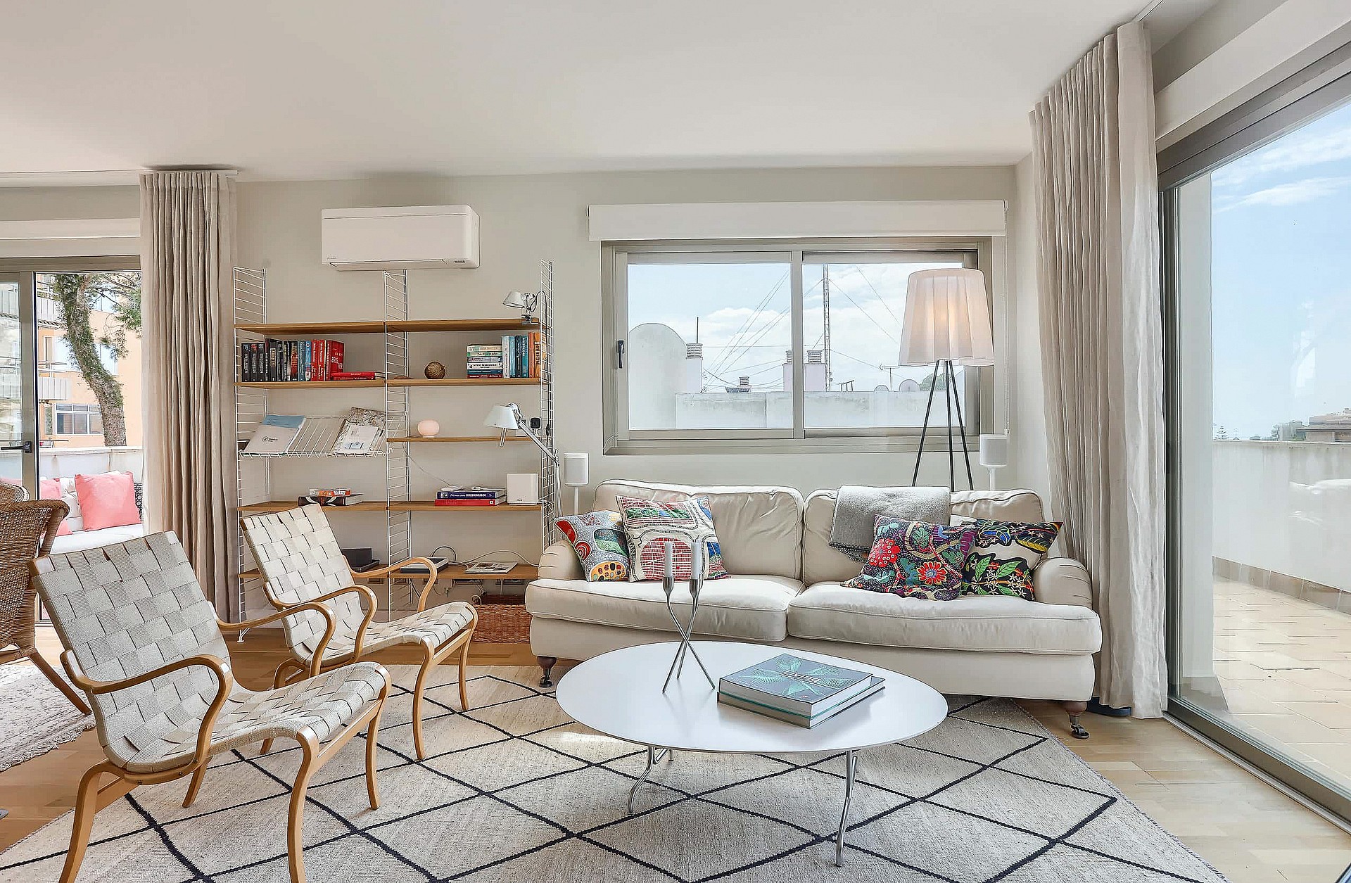 Bright living room featuring a neutral sofa, patterned armchairs, metal shelving, and a large window leading to a terrace.