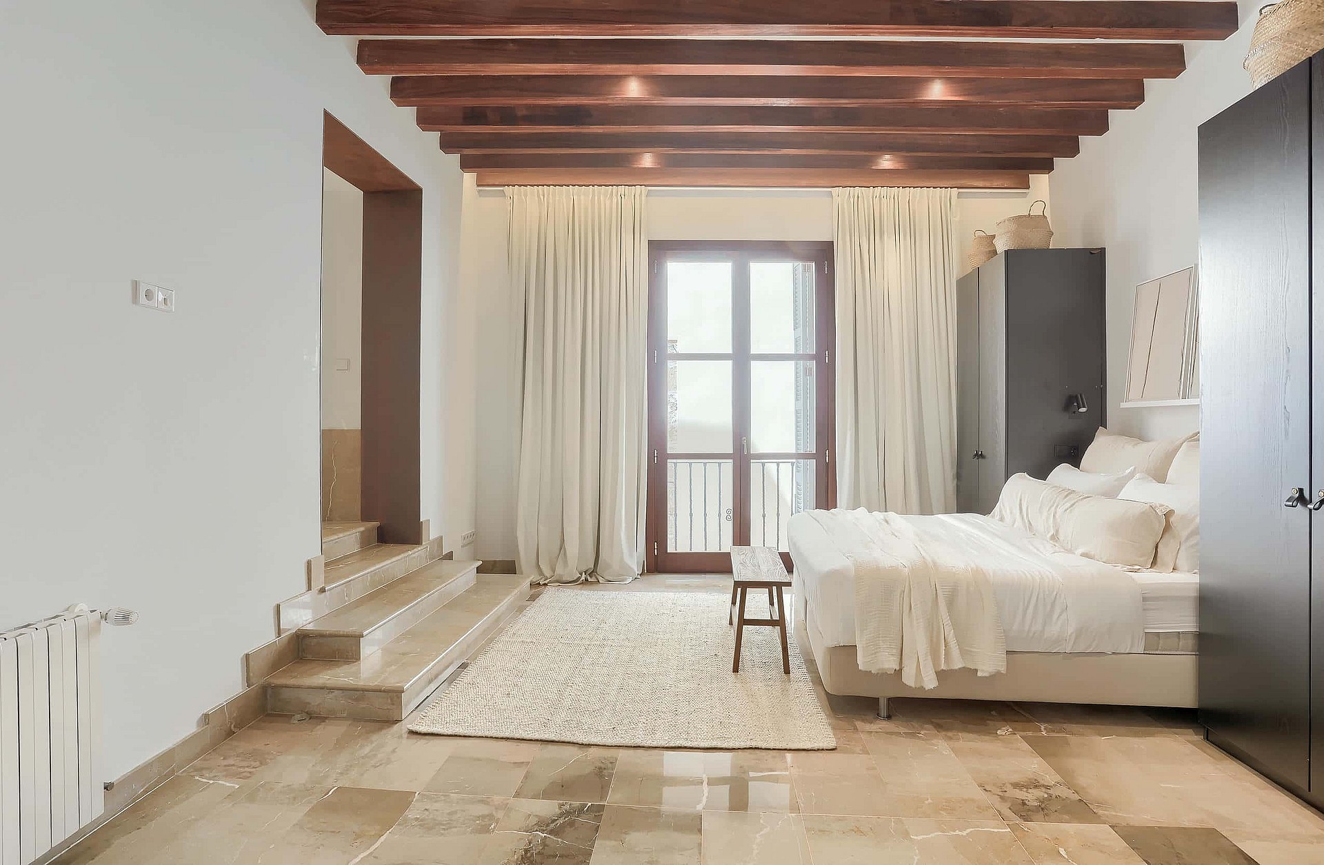 Spacious bedroom with exposed wood ceiling beams, stone flooring, a double bed, and glass doors leading to a balcony.