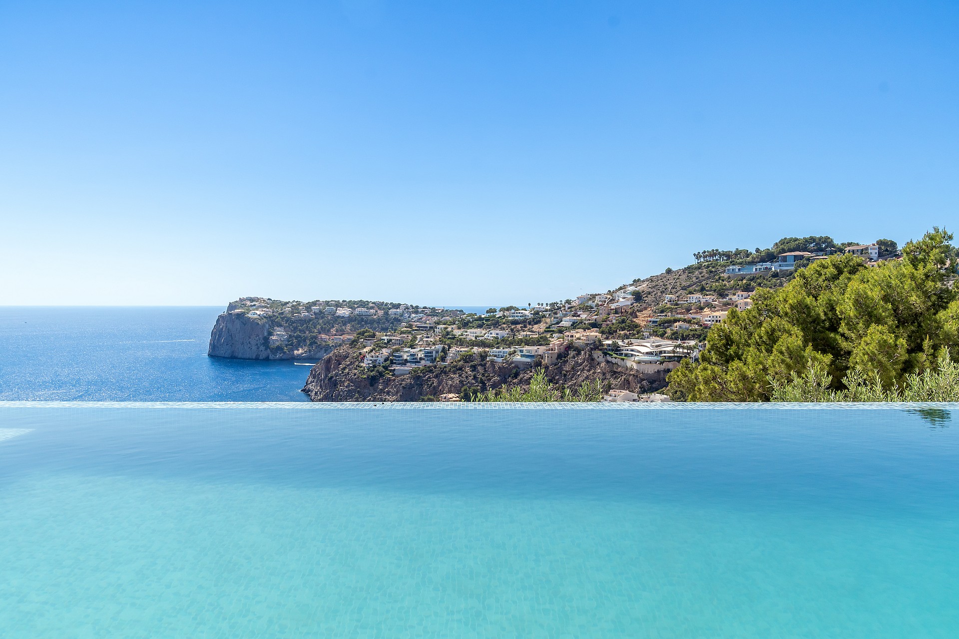 A clear blue infinity pool edge meets the horizon, overlooking a sunlit hillside with coastal villas and green trees.
