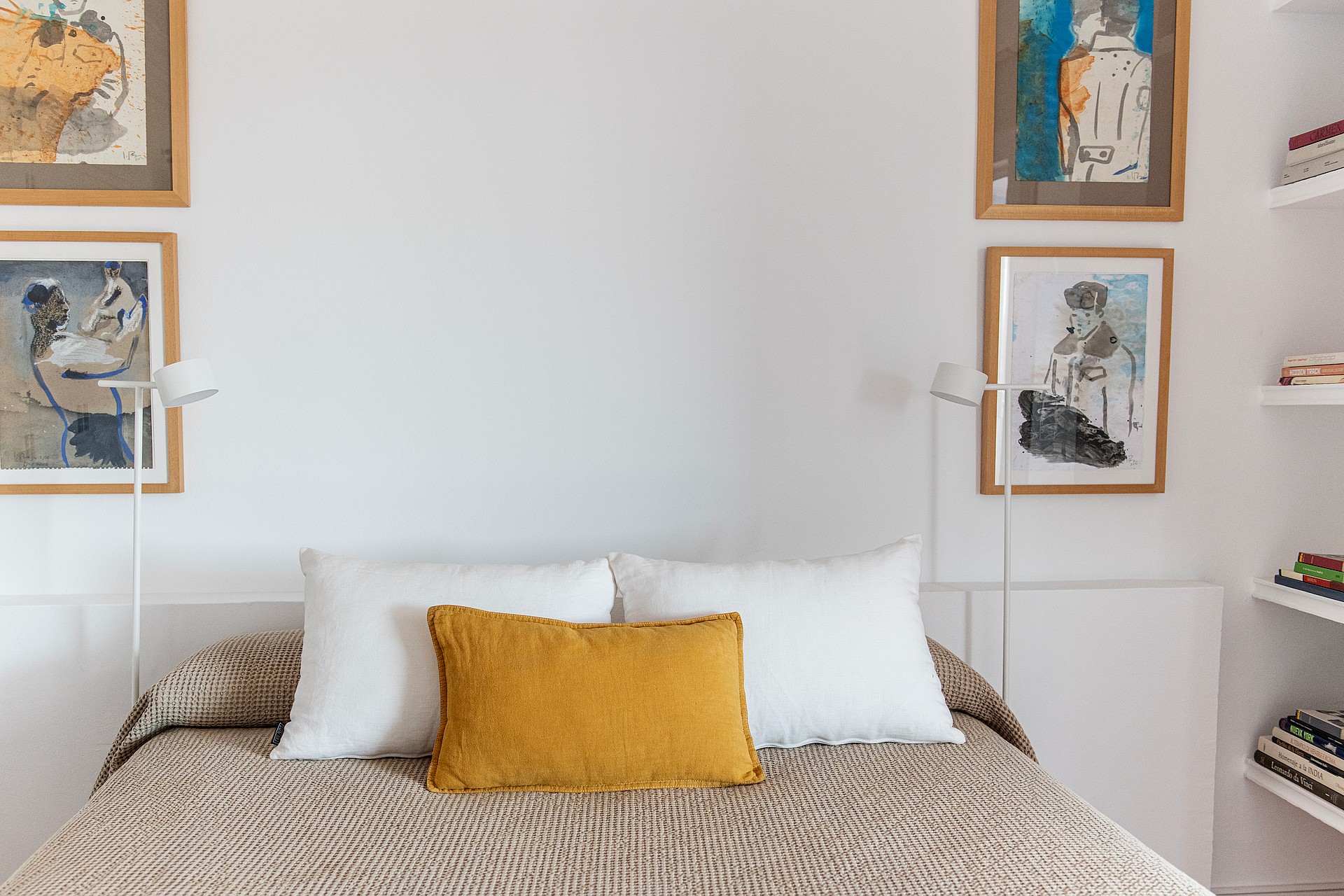 A double bed with a patterned beige cover and mustard yellow cushion, flanked by two minimalist floor lamps and framed wall art.