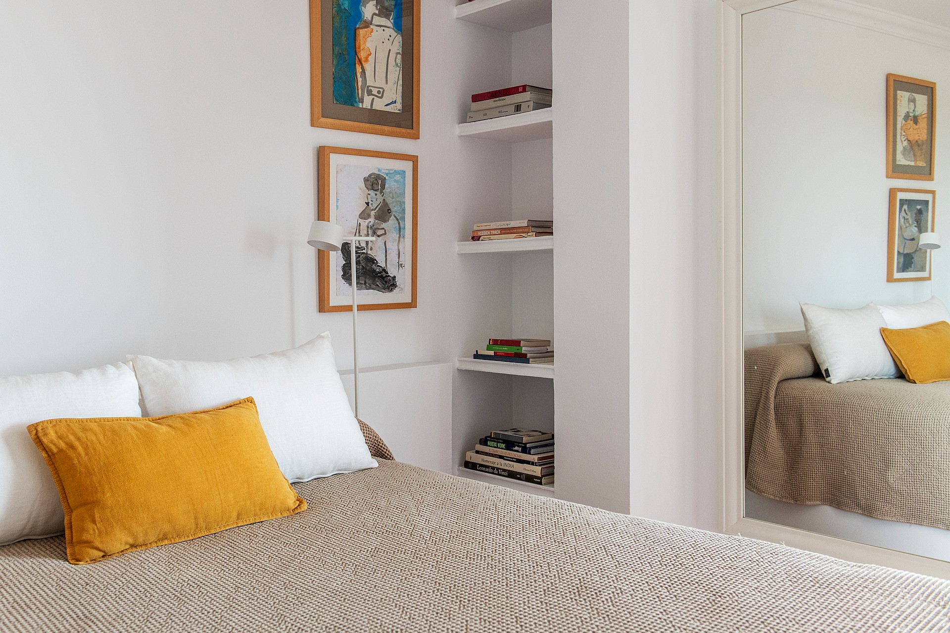 A close-up of a bedroom featuring a textured beige bedspread, a bright yellow throw pillow, and integrated white wall shelving.