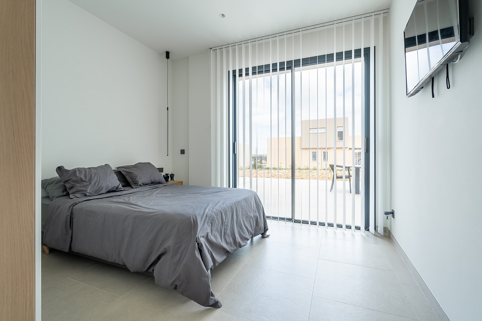 Modern bedroom with a grey bed, concrete floor, and vertical blinds covering a glass door to a terrace.