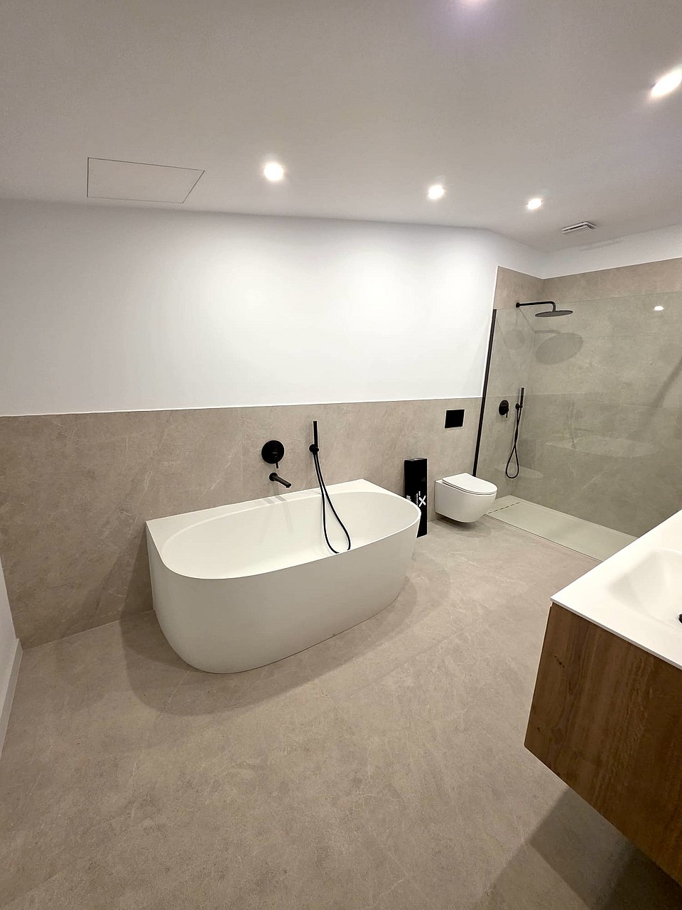 A modern bathroom featuring a white freestanding bathtub, beige stone floor tiles, a walk-in shower, and black fixtures.