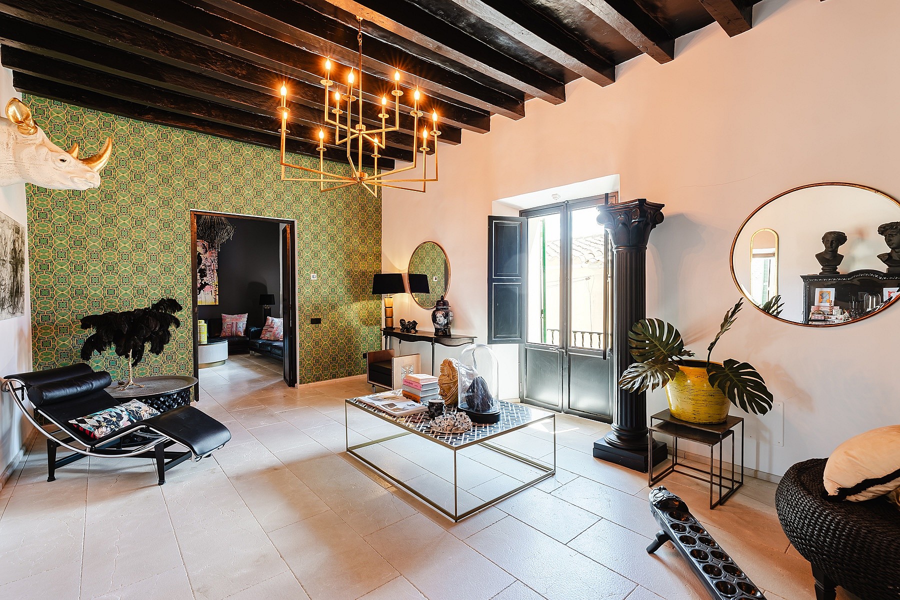 A spacious living room featuring a dark wooden beamed ceiling, a patterned green accent wall, and a large metal chandelier.