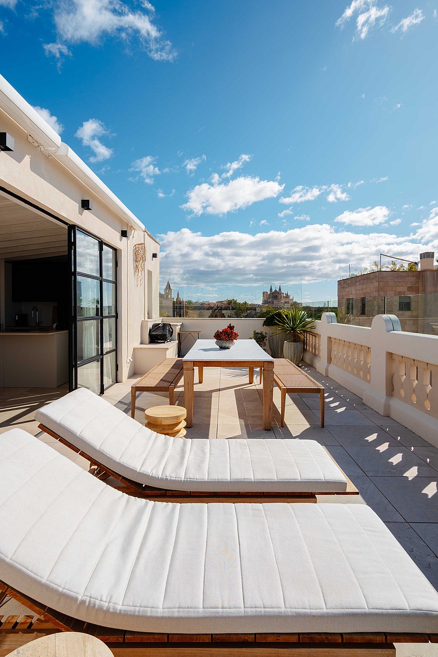 An expansive outdoor rooftop terrace featuring white sun loungers, a wooden dining table, and a view over the Santa Catalina skyline under a clear blue sky.