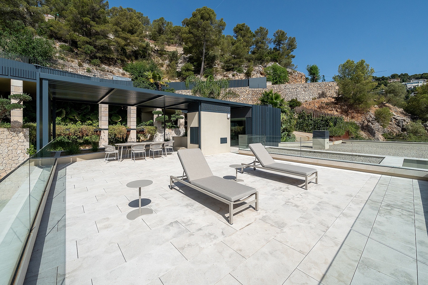 A wide outdoor terrace with stone paving, two sun loungers, and a covered dining area set against a hillside backdrop.