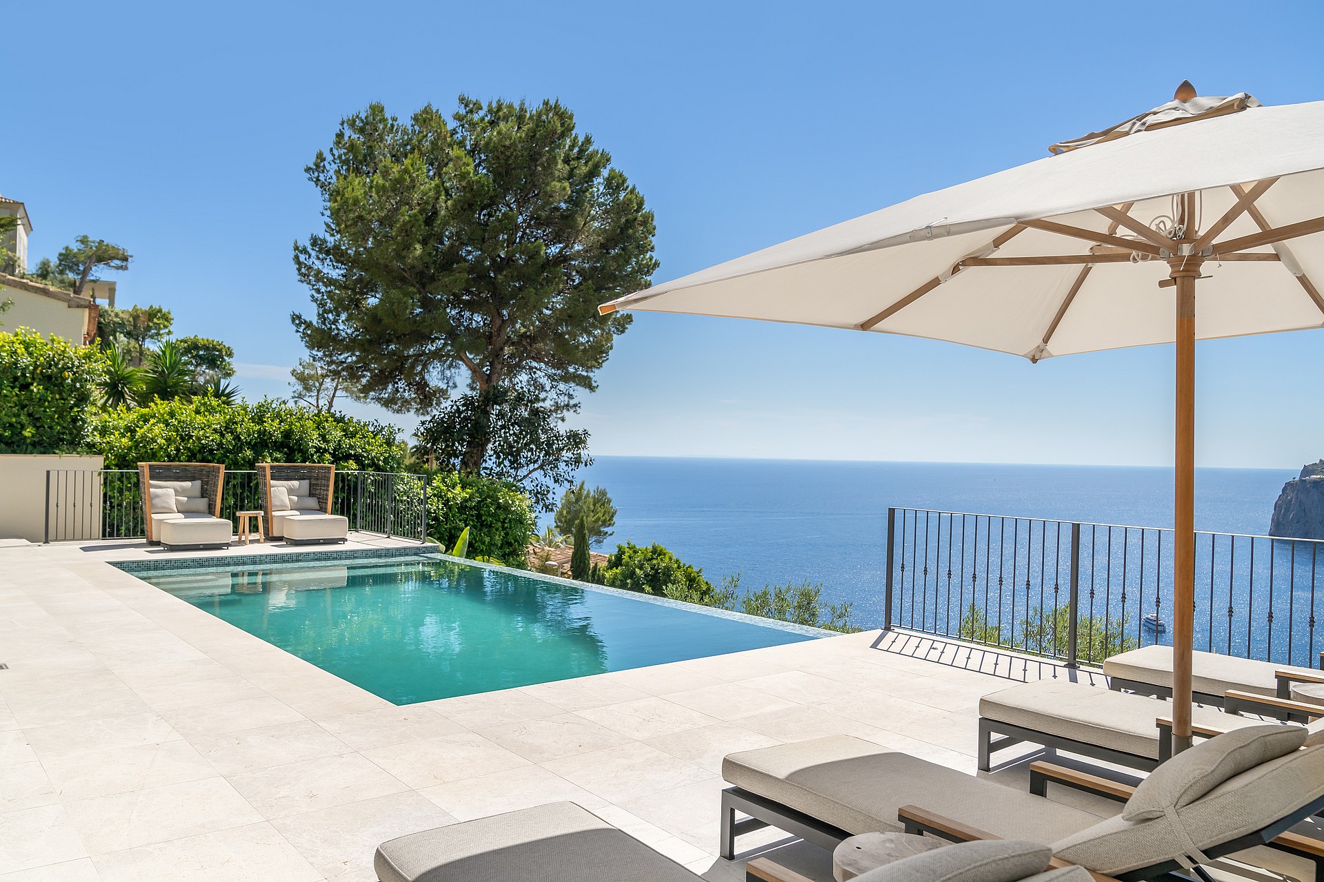 A rectangular swimming pool on a pale stone terrace overlooks the blue sea, framed by a pine tree and lounge furniture.