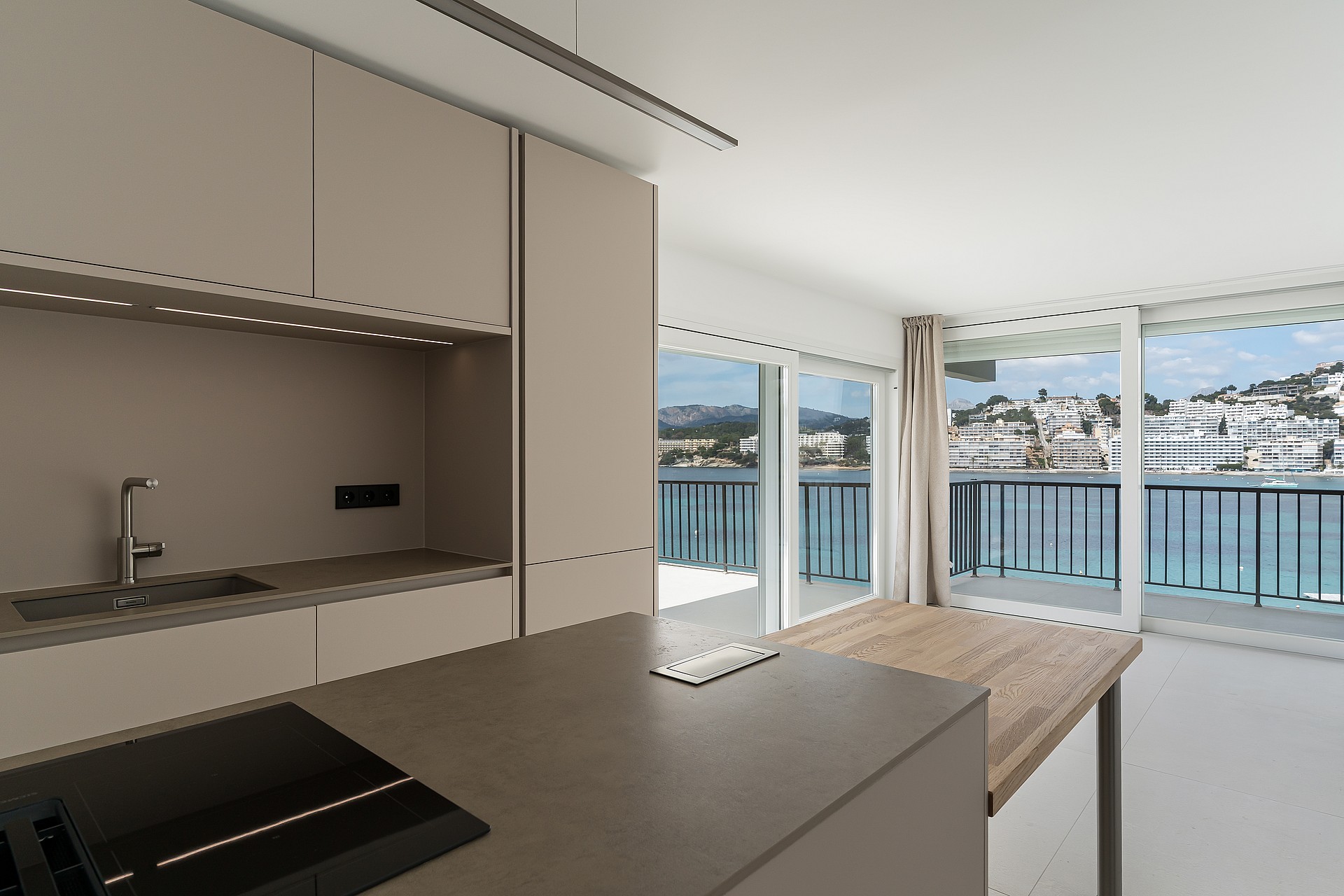 Modern taupe kitchen and wooden island extension facing floor-to-ceiling windows with a view of a coastal harbor.