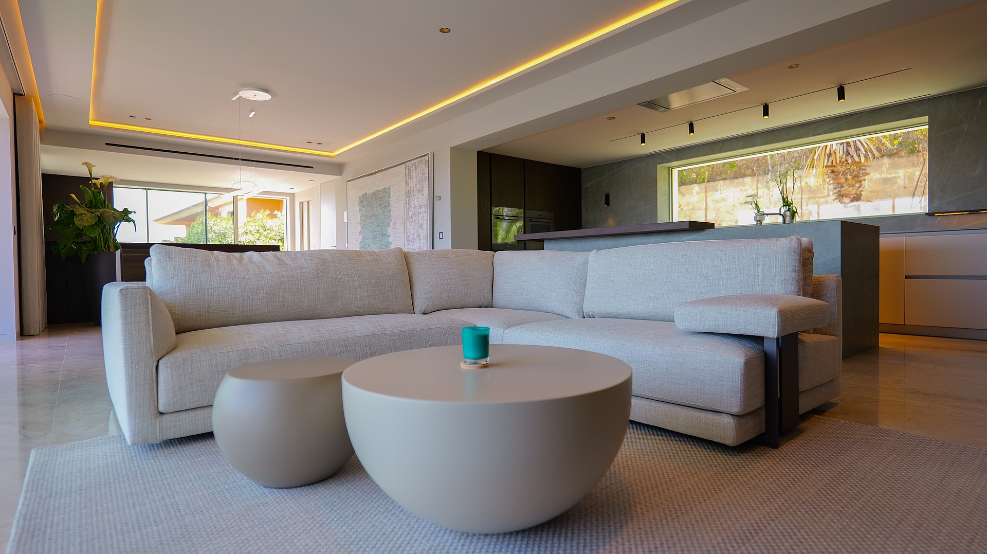A contemporary open-plan living room featuring a light grey modular sofa, a rounded coffee table, and an integrated kitchen with a dark stone island and horizontal window.