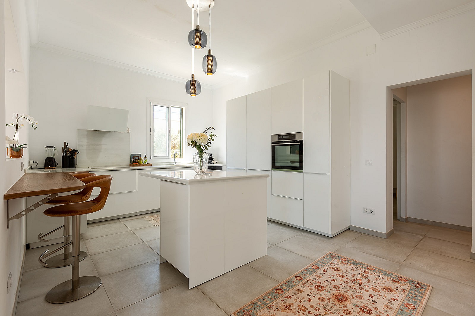 Modern white kitchen featuring a central island, integrated appliances, bar seating, and a patterned area rug on tiled floors.