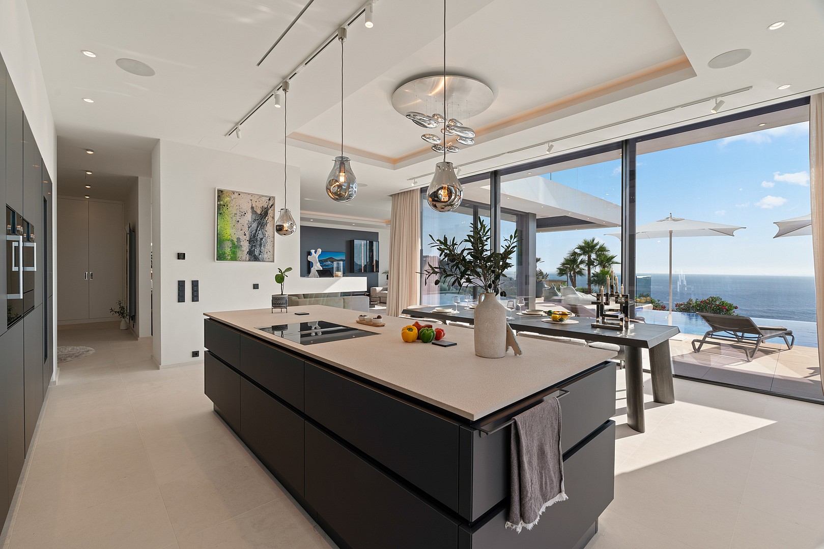 Modern open-plan kitchen with a large dark charcoal island, light stone countertop, and floor-to-ceiling glass doors opening to a sea-view terrace.