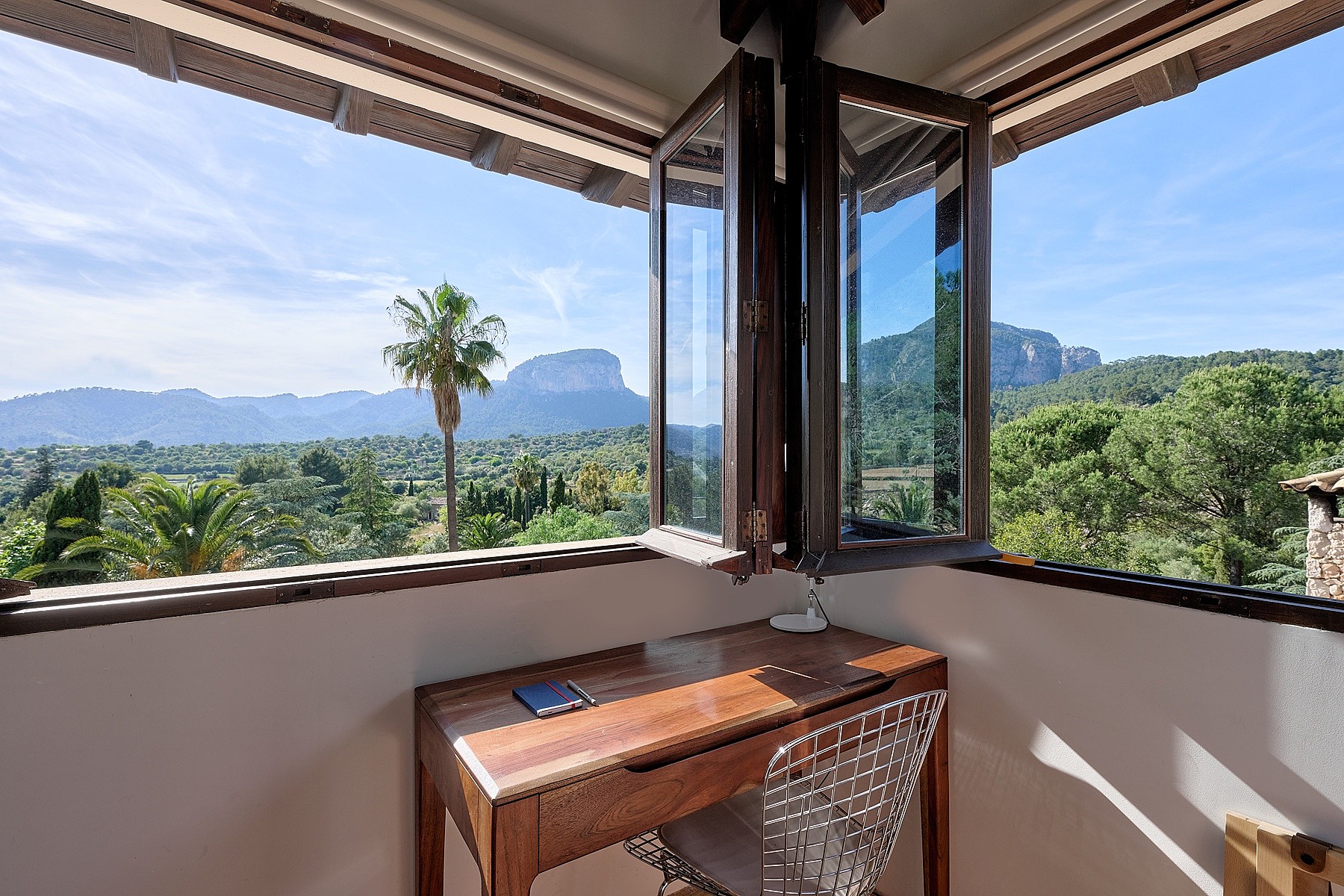 A small wooden desk positioned in a corner with floor-to-ceiling windows framing a landscape of Alaro mountains.
