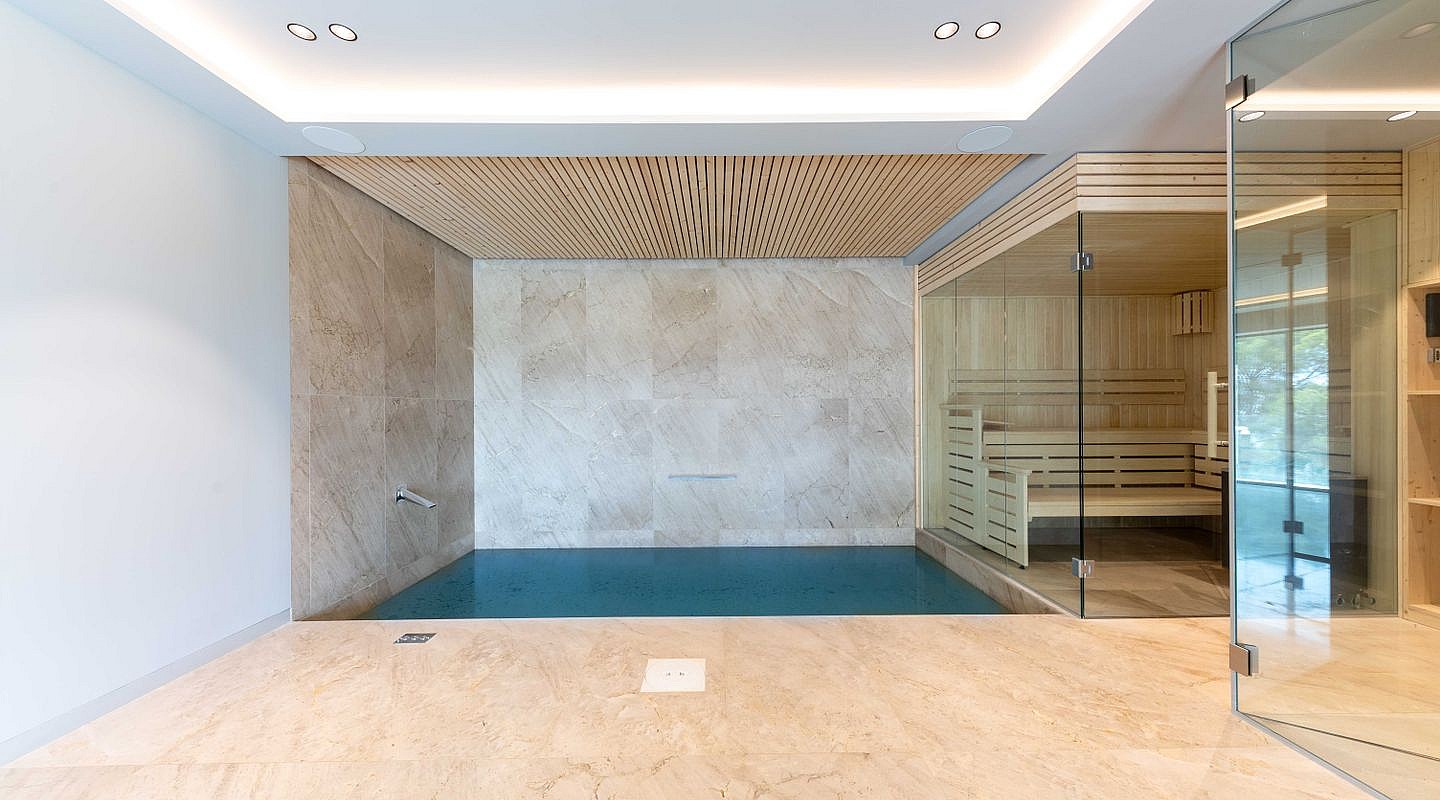 Indoor pool and sauna area with light wood slatted ceiling, large stone floor tiles, and glass partitions.