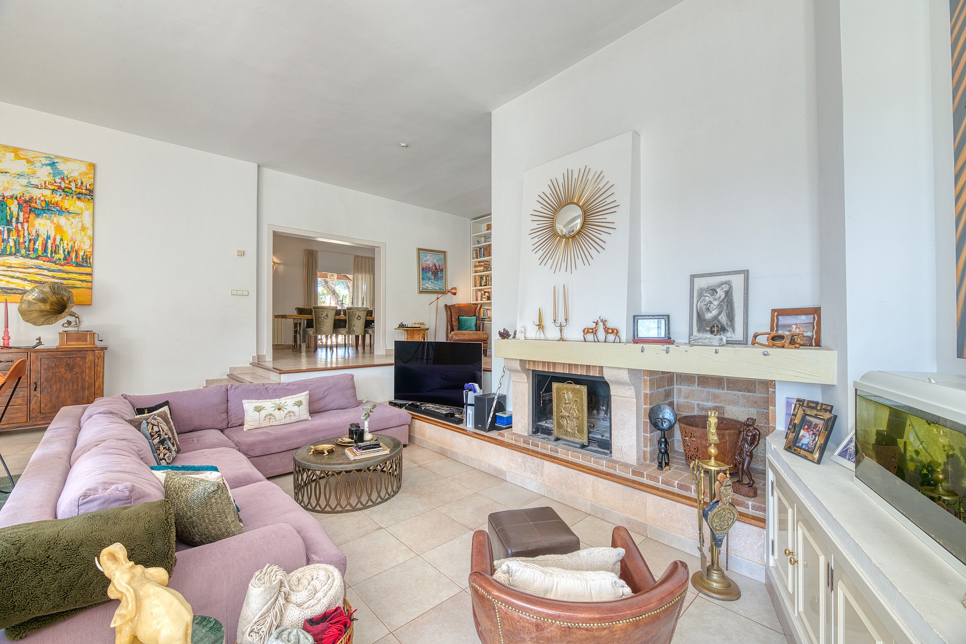Spacious living room featuring a large lilac sectional sofa, a stone-fronted fireplace, and a light-colored tiled floor.