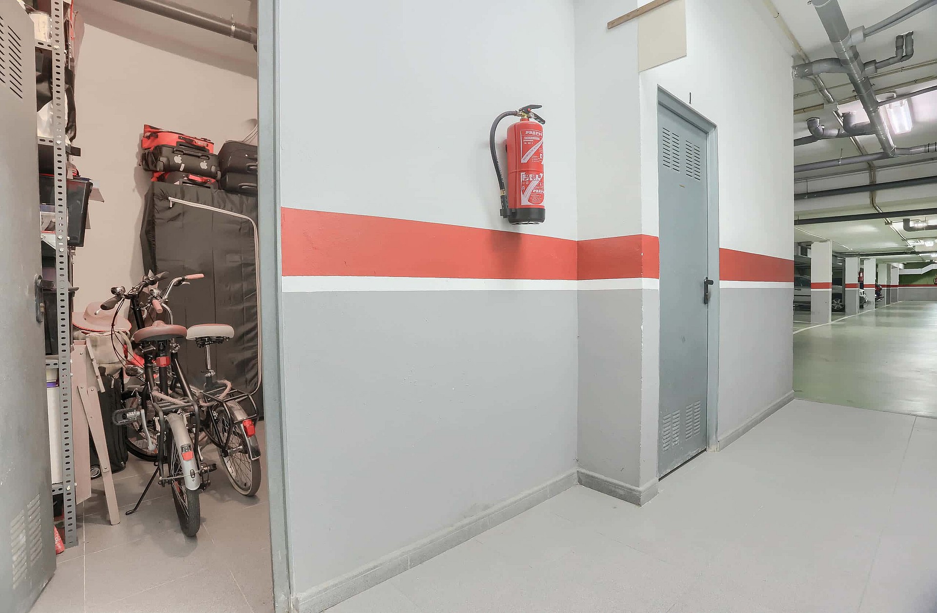 A private storage room with bicycles, located next to a grey painted wall with a fire extinguisher inside an underground parking garage.