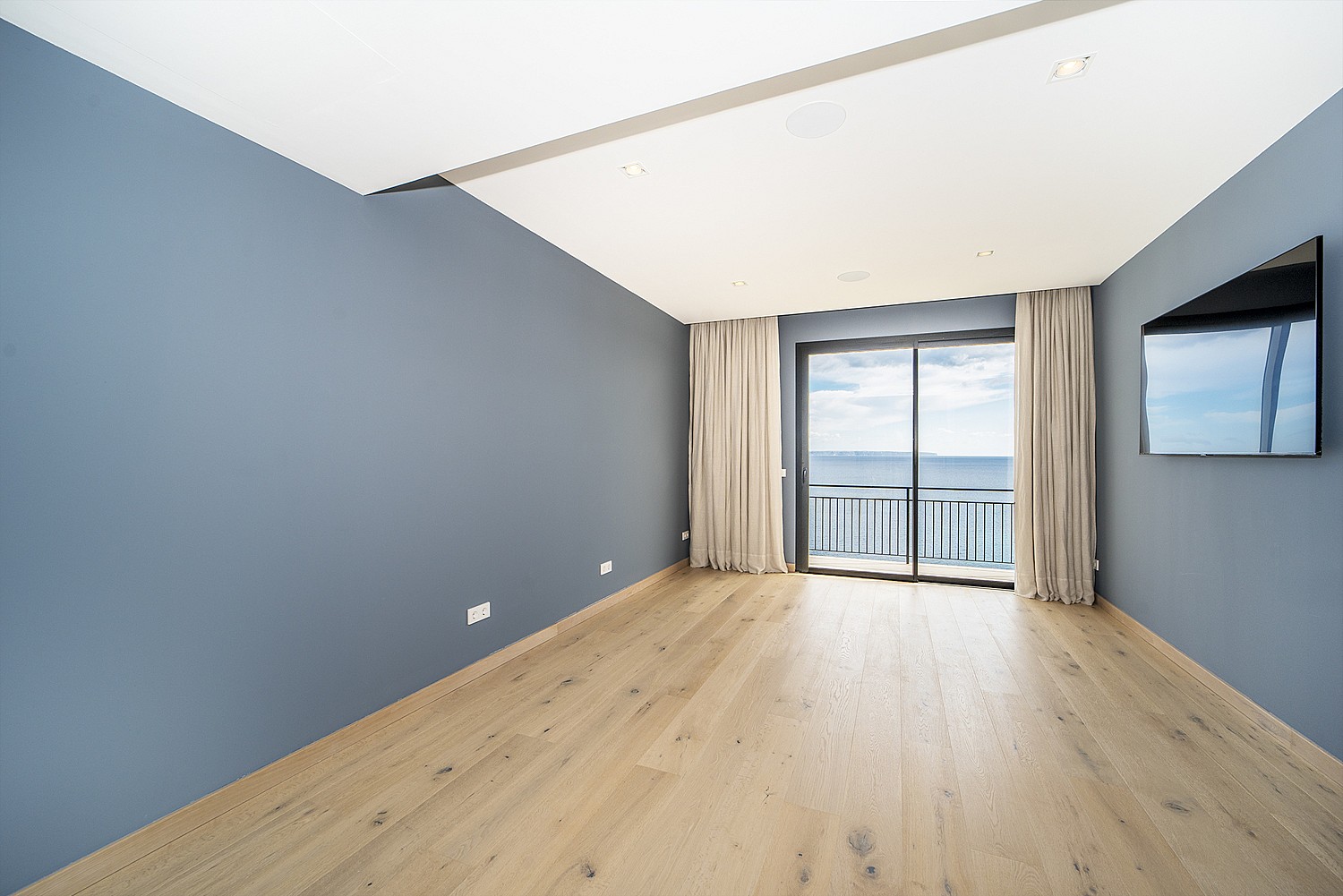 An empty bedroom with blue walls, light oak wood floors, and large glass sliding doors opening to a balcony with sea views.
