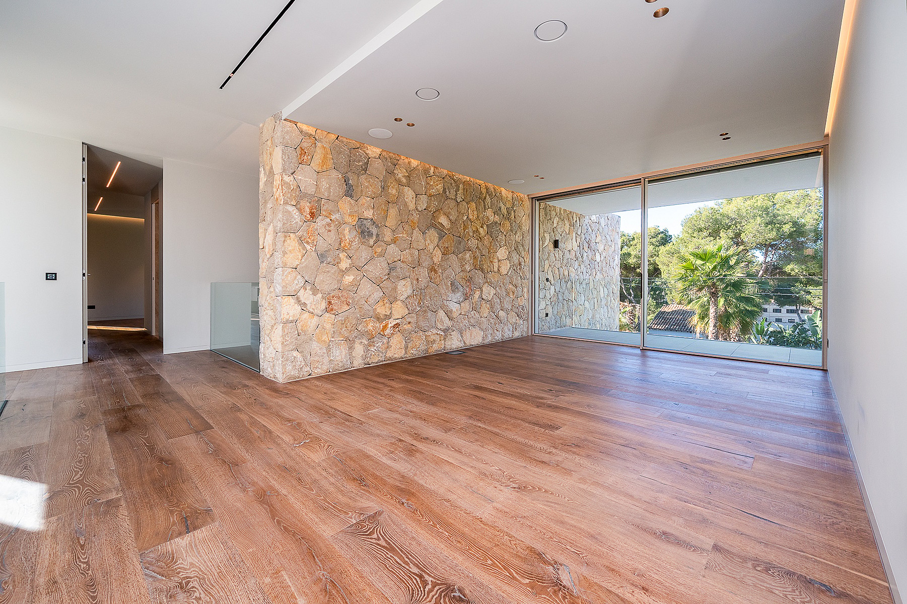 Empty modern living area with light oak floors and a large dry-stone accent wall facing floor-to-ceiling windows.