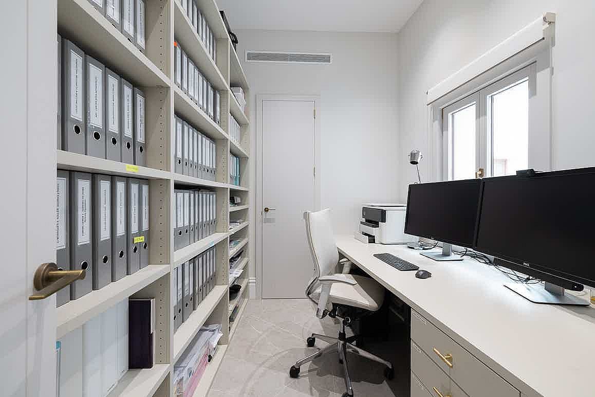 A compact home office featuring floor-to-ceiling built-in shelving packed with grey binders, a long desk with dual monitors, and a white task chair.