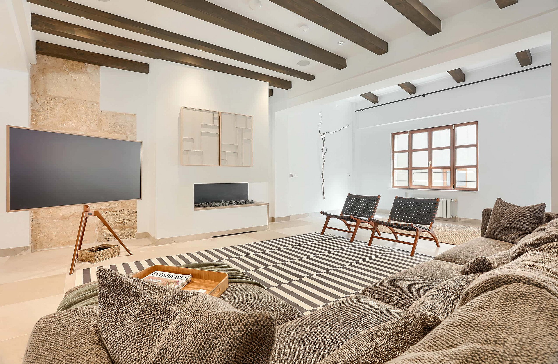 A living room interior featuring dark wood exposed ceiling beams, a textured stone wall, a large flat-screen TV on a wooden tripod stand, and two mid-century style armchairs on a patterned rug.