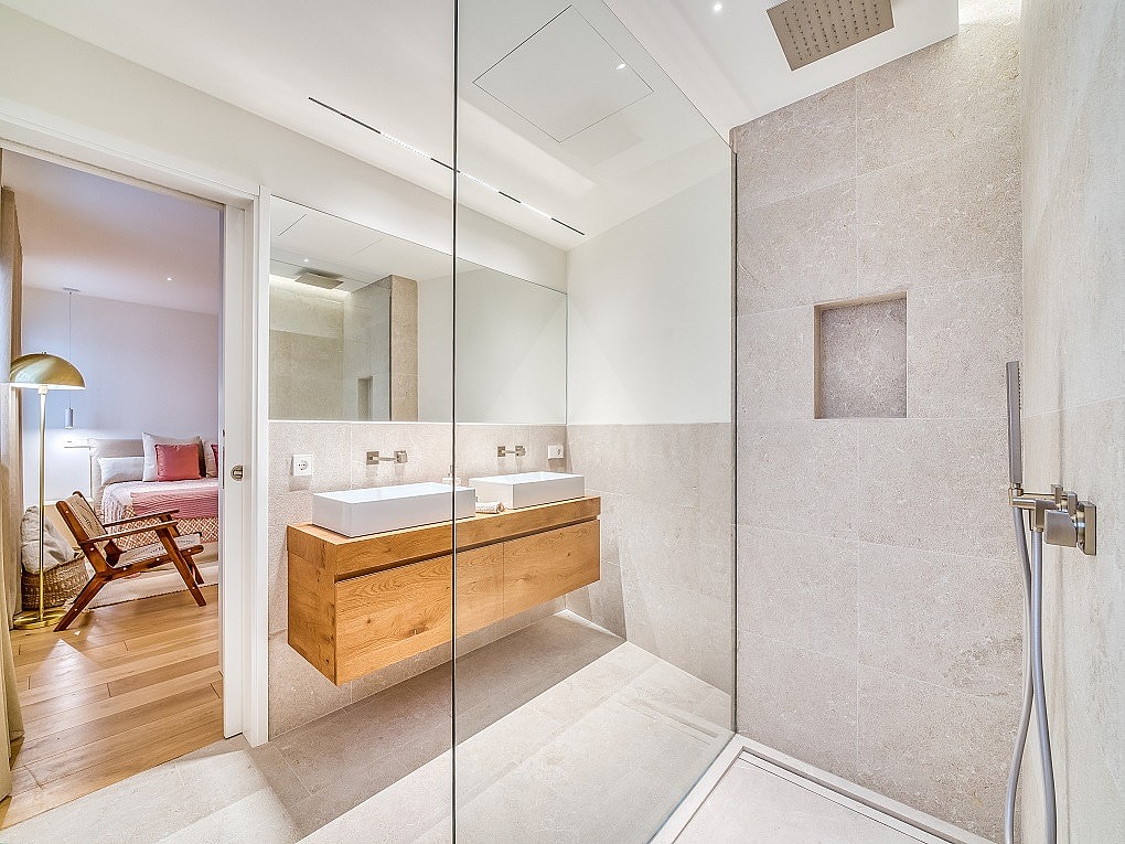 Interior view of a contemporary bathroom featuring a floating light oak vanity, dual ceramic basins, and a glass-enclosed shower with textured stone tiles.