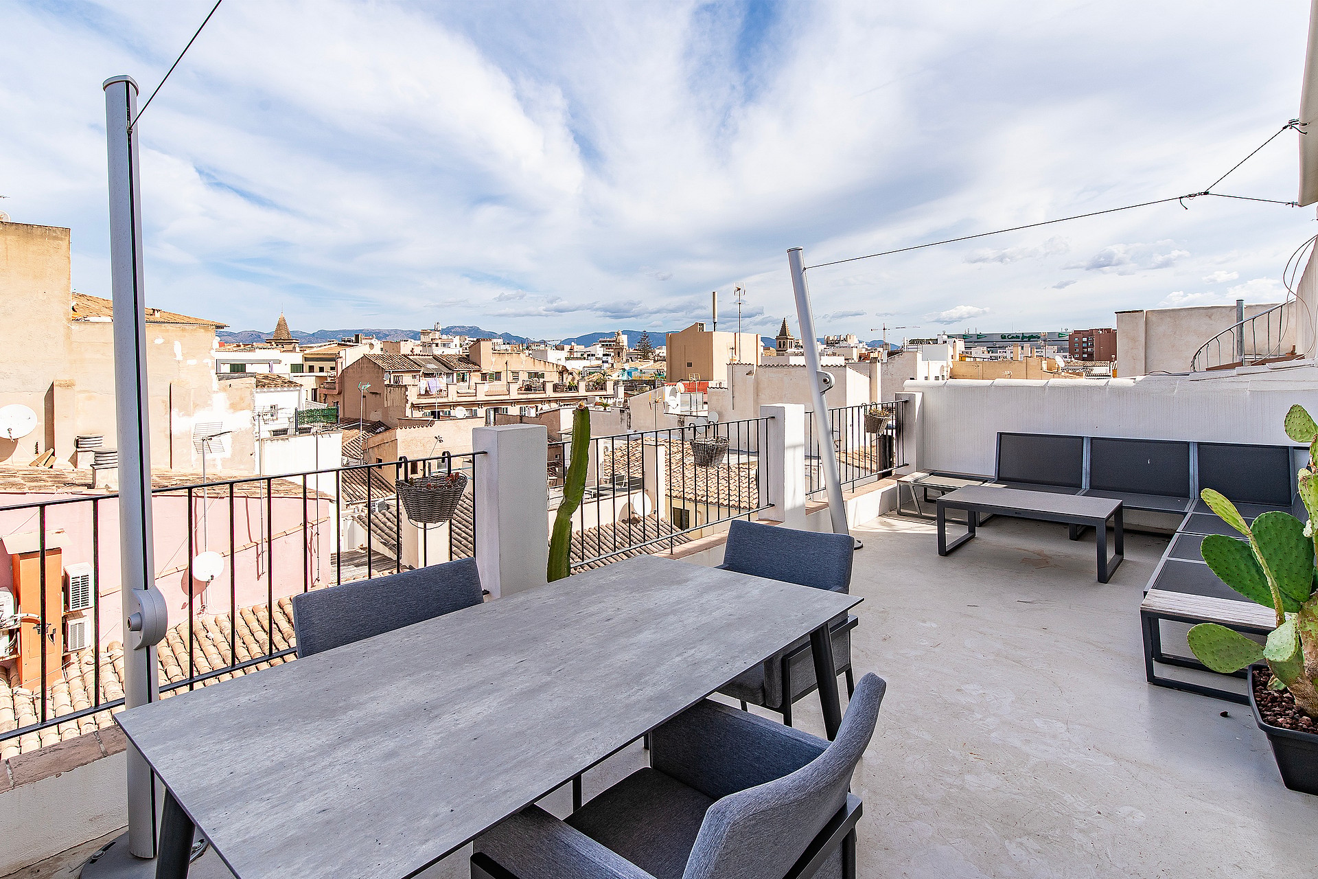 An outdoor terrace in an urban setting featuring a dining table, grey upholstered seating, and panoramic views of old town rooftops under a blue sky.