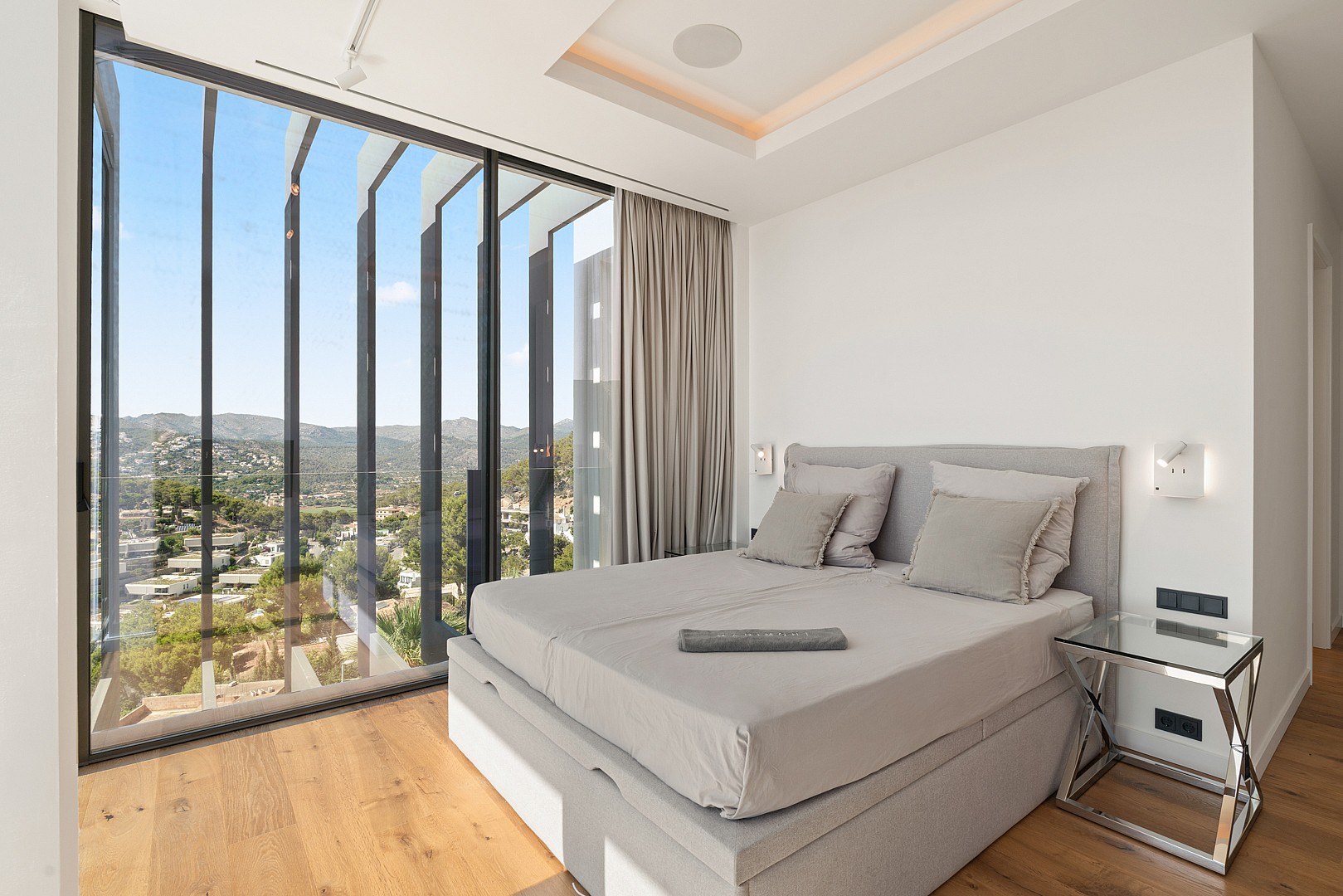 Corner bedroom with floor-to-ceiling windows looking out over a coastal landscape, featuring a grey bed and oak flooring.