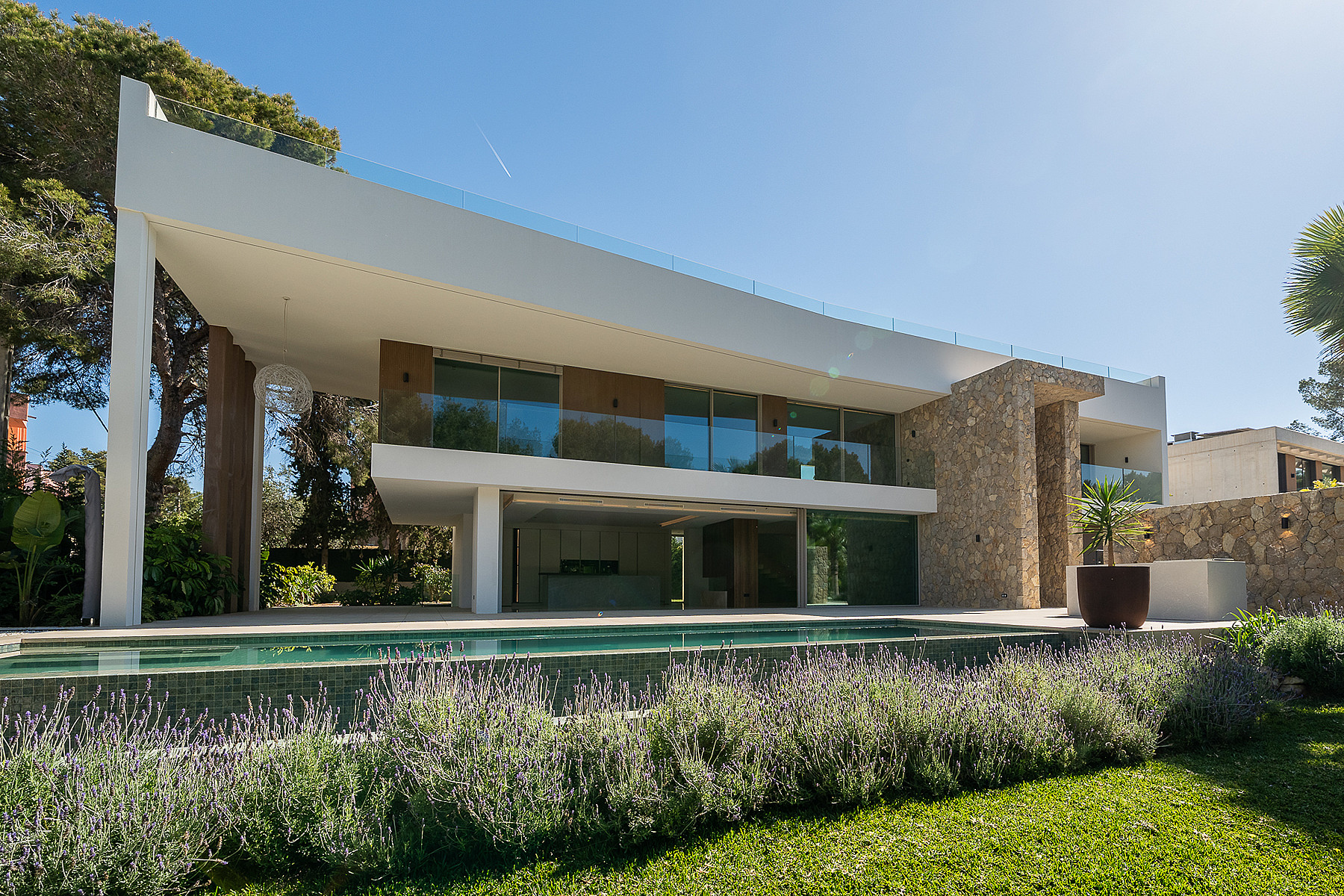 Exterior view of a two-story modern villa featuring a stone wall, glass railings, and a swimming pool in the foreground.
