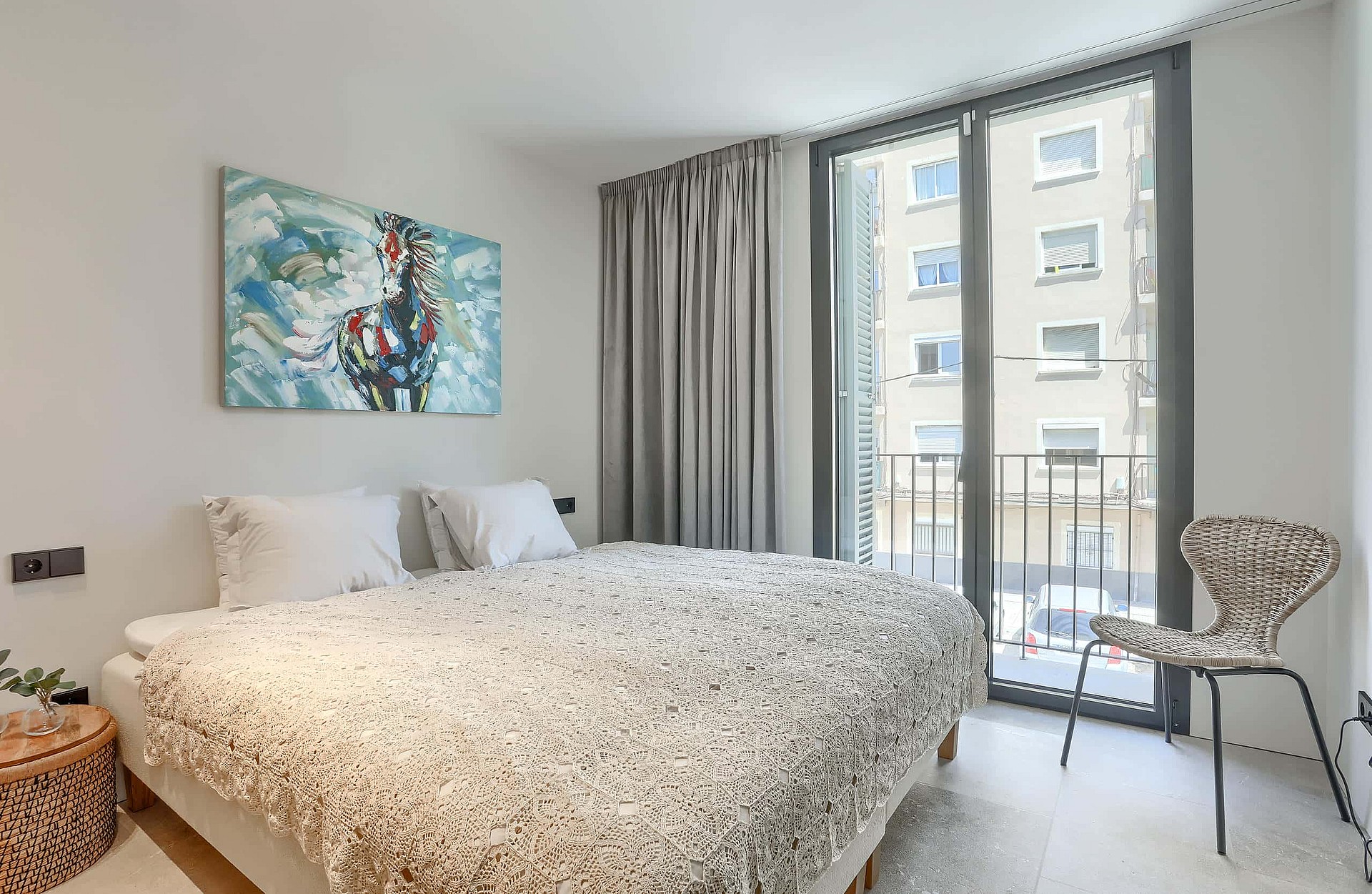 A contemporary bedroom featuring a double bed, patterned bedding, grey curtains, and a Juliet balcony overlooking a street.