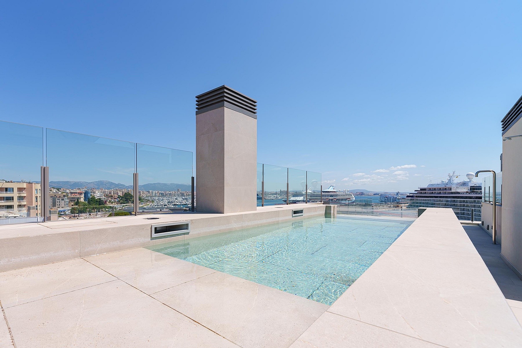 Piscina en azotea con barandillas de cristal hacia el mar, con pavimento de piedra y chimenea central.
