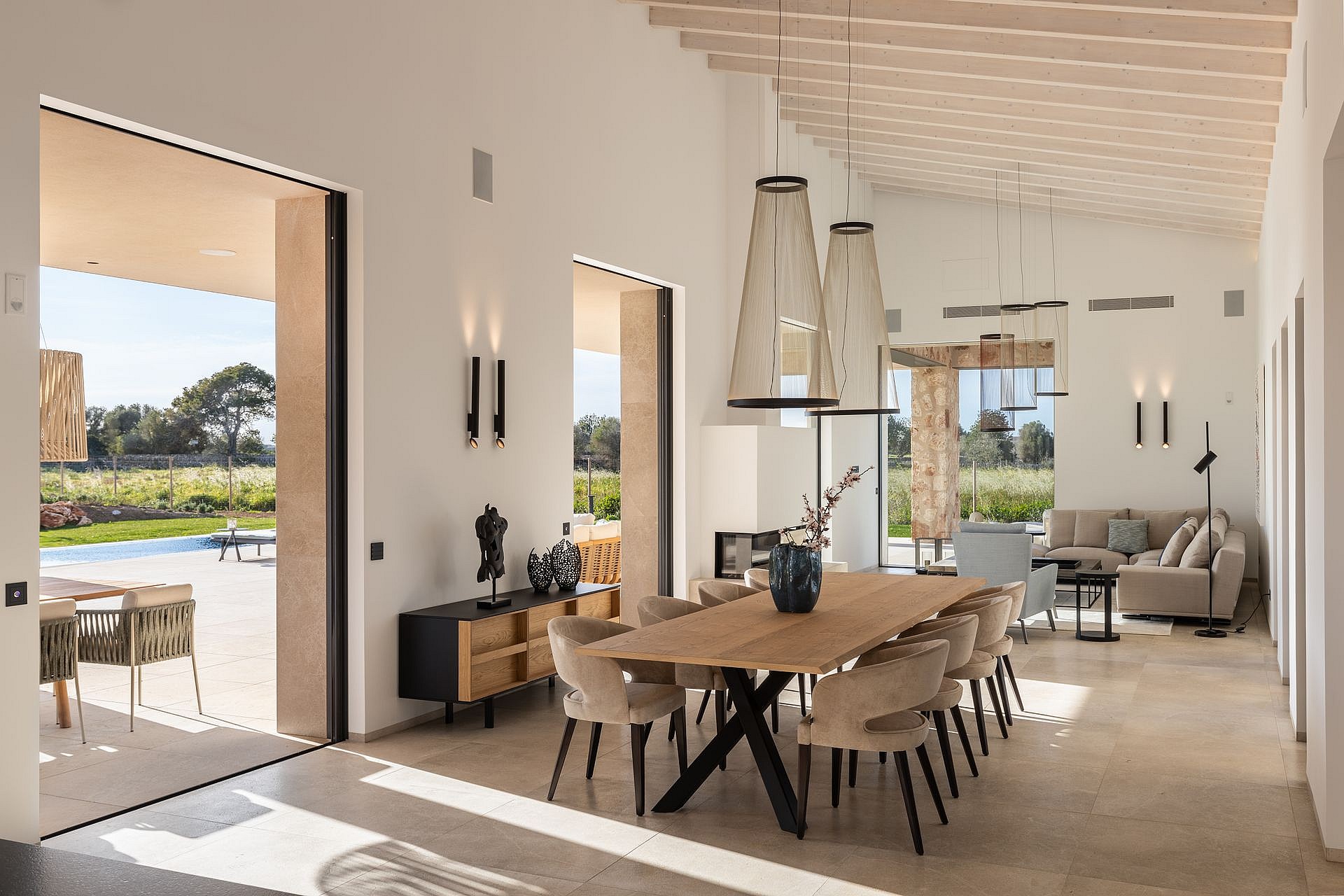 Dining room with a long oak table and cushioned chairs, open to an outdoor terrace, with white walls and exposed ceiling beams.