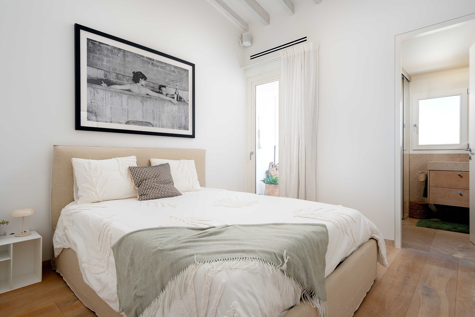 A bright bedroom with a queen-sized bed, white linens, a neutral headboard, framed black and white art, and a view into an en-suite bathroom.