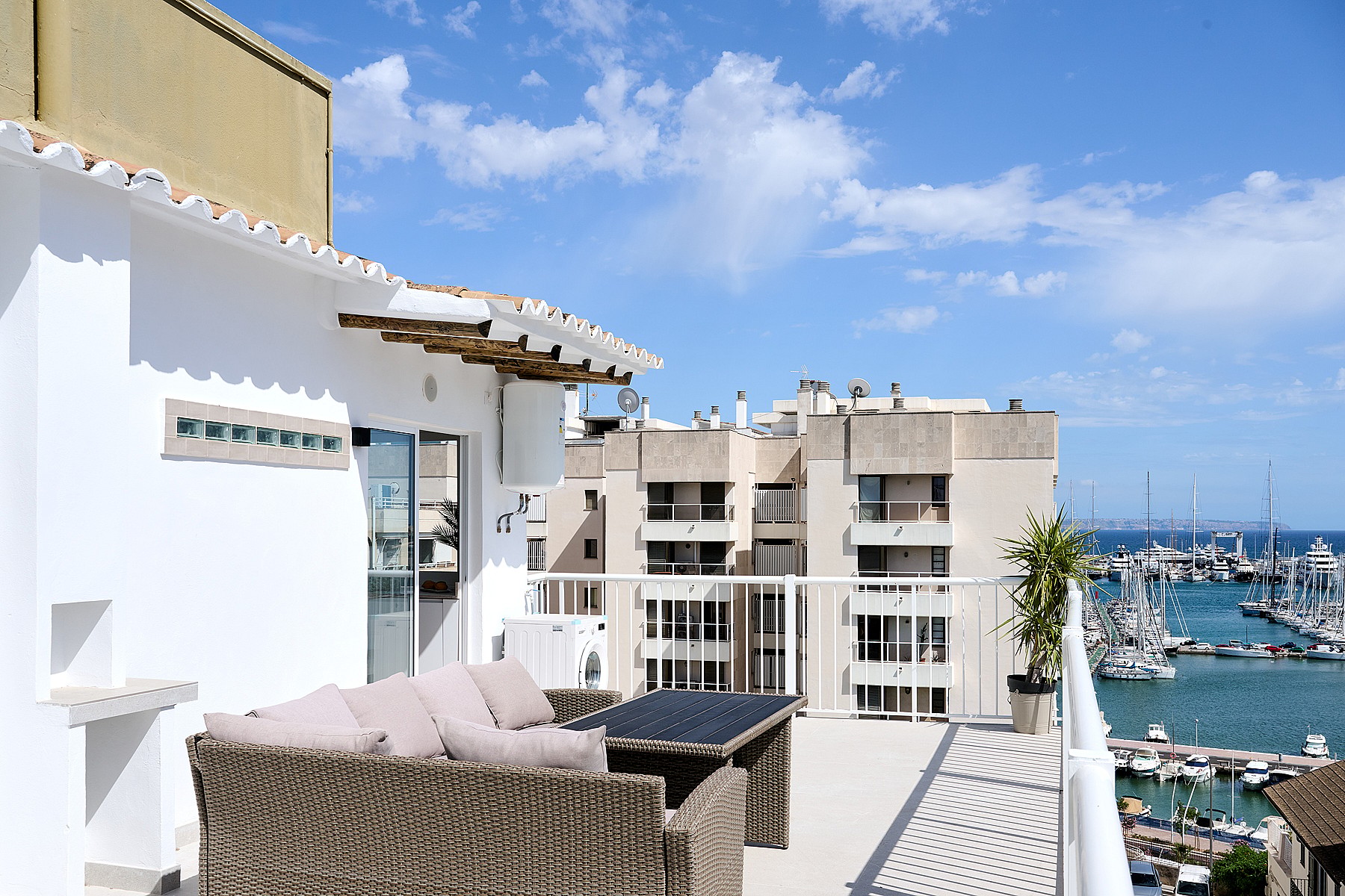 A rooftop terrace with rattan seating, overlooking a marina with boats and nearby apartment buildings under a blue sky.