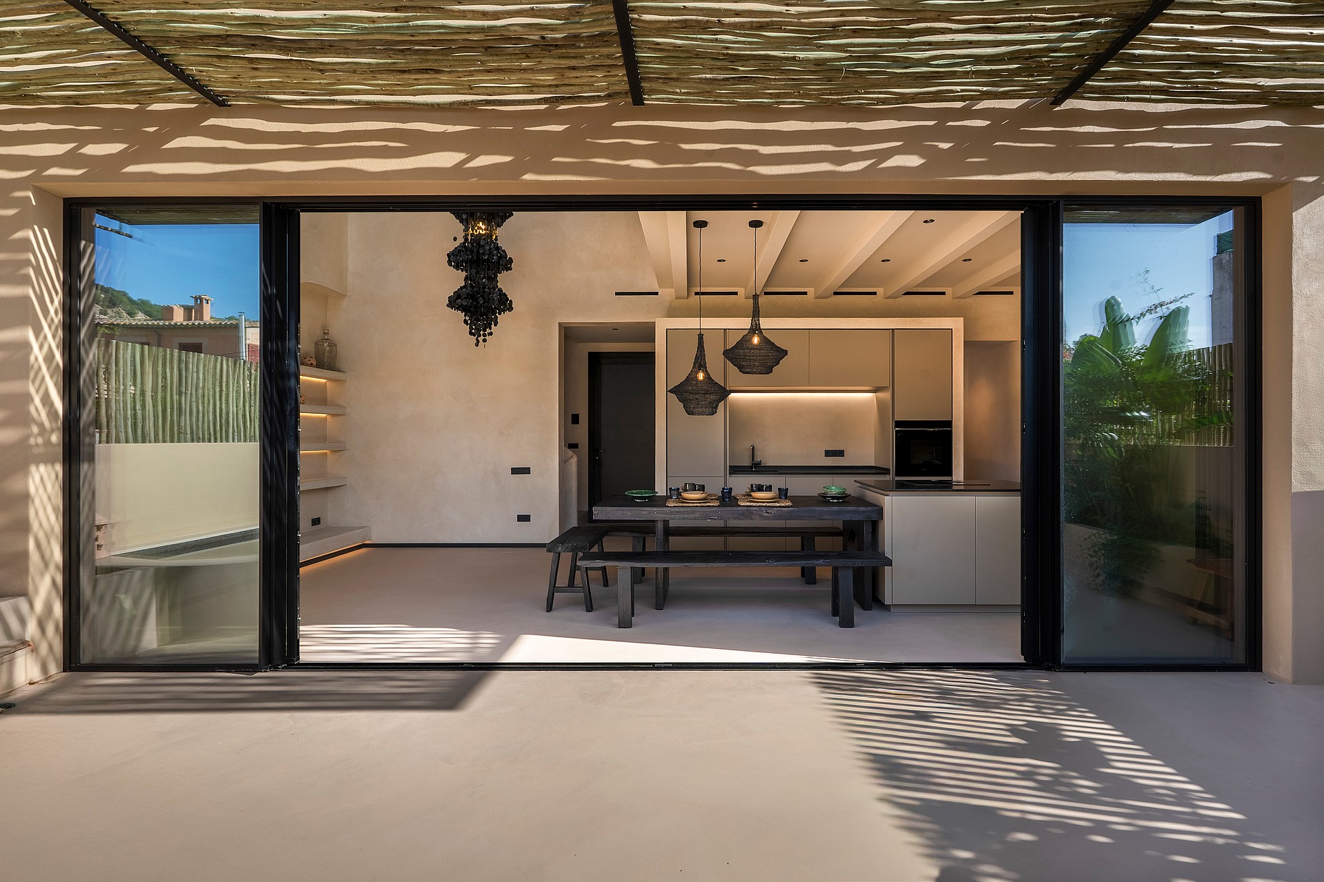 A modern kitchen and dining space viewed from a terrace through large sliding glass doors, featuring plastered walls and wooden ceiling beams.