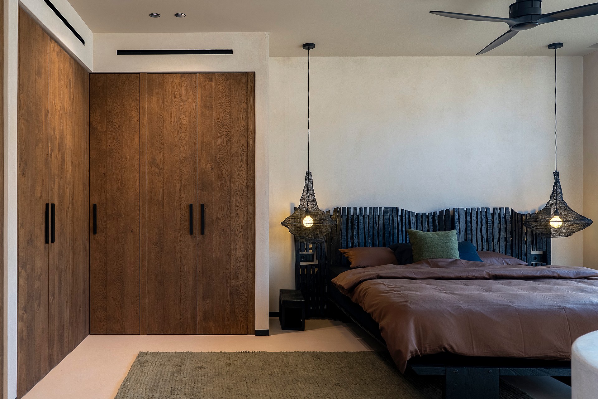 A modern bedroom featuring dark wood built-in wardrobes, a bed with a charred timber headboard, and pendant lights.